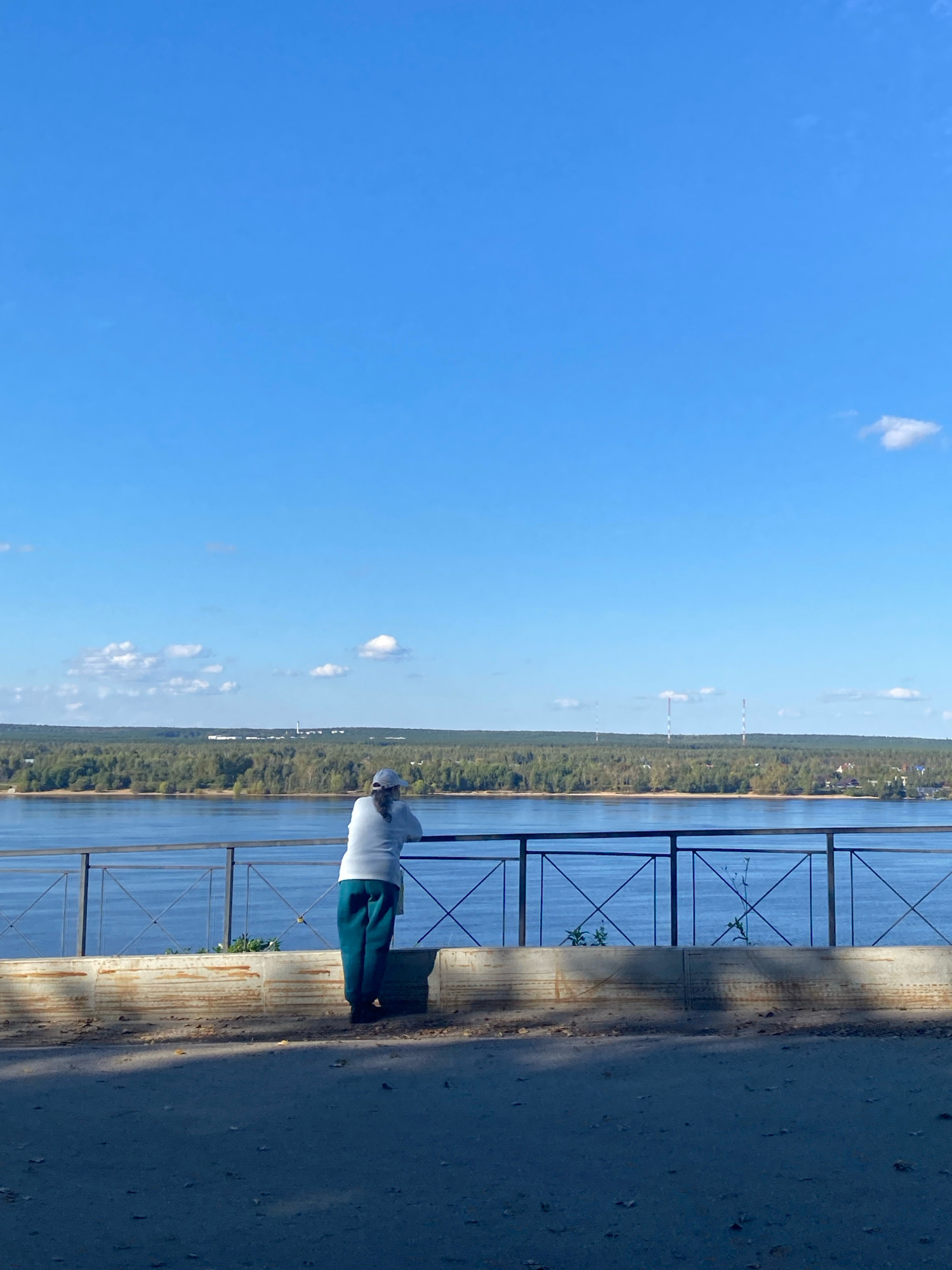 An individual stands facing a body of water, leaning on a black metal railing. The person is wearing a white long-sleeved shirt, a light-colored cap, and teal pants. Behind the railing is a wide river or bay, with a treelined shore on the opposite bank. The sky is clear and blue with scattered white clouds. The foreground shows a paved area, possibly a sidewalk or walkway, with some fallen leaves. In the distance across the water, several tall, thin structures, possibly industrial smokestacks or radio towers, are visible. This scene is likely along the Kama River embankment in the city of Perm, Russia, a popular spot for river views.
