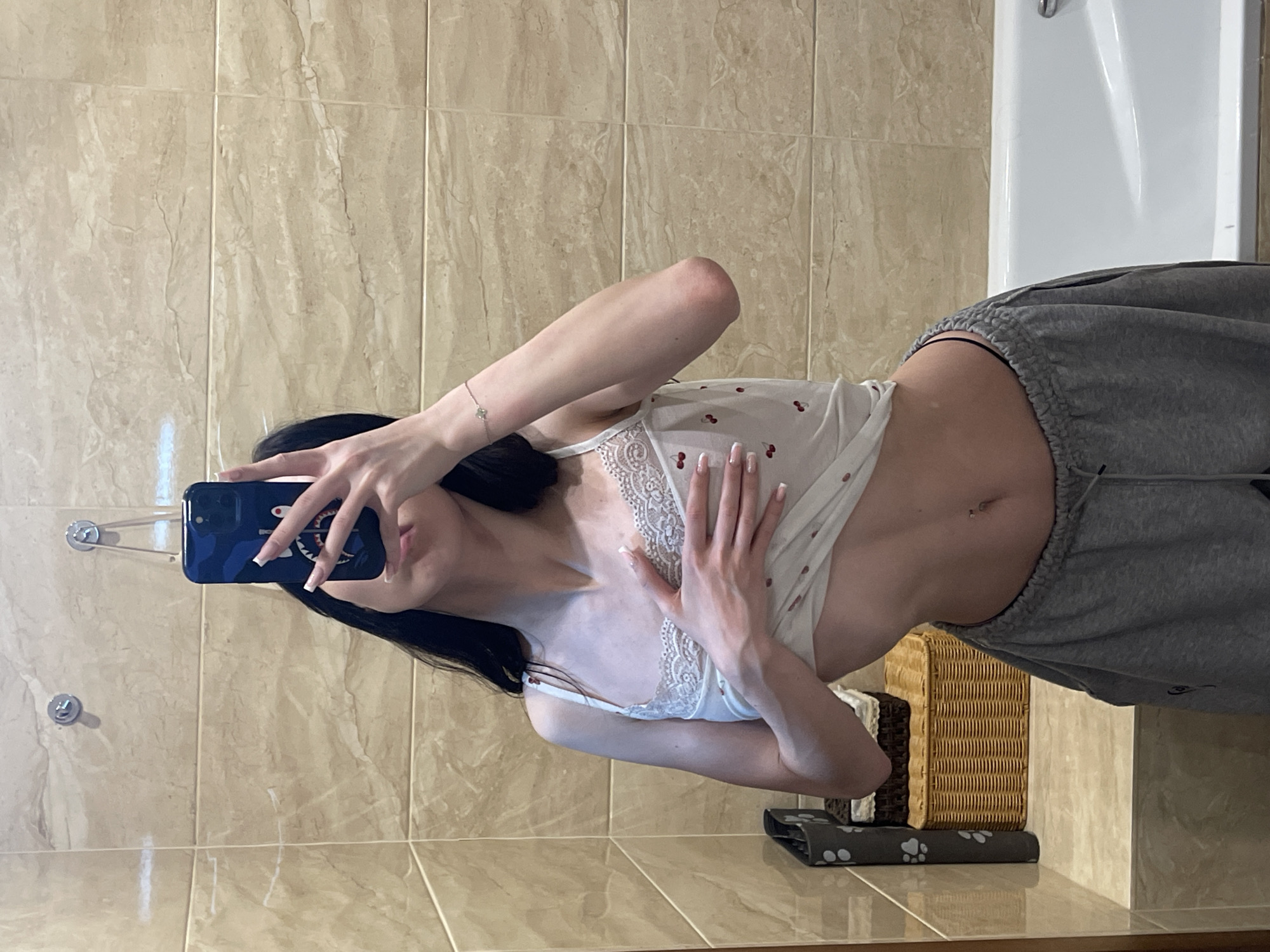 A young woman with long dark hair takes a selfie in a bathroom. She is wearing a white camisole with lace trim and a cherry pattern, and grey sweatpants. Her left hand is on her chest, and her right arm is bent at the elbow. The bathroom has beige tiled walls and a white bathtub. A wicker basket is visible on a shelf to the left of the woman, next to a grey laundry bin with paw prints. The scene is likely indoors, in a residential setting, with no visible indication of the specific city of Khimki, Russia.