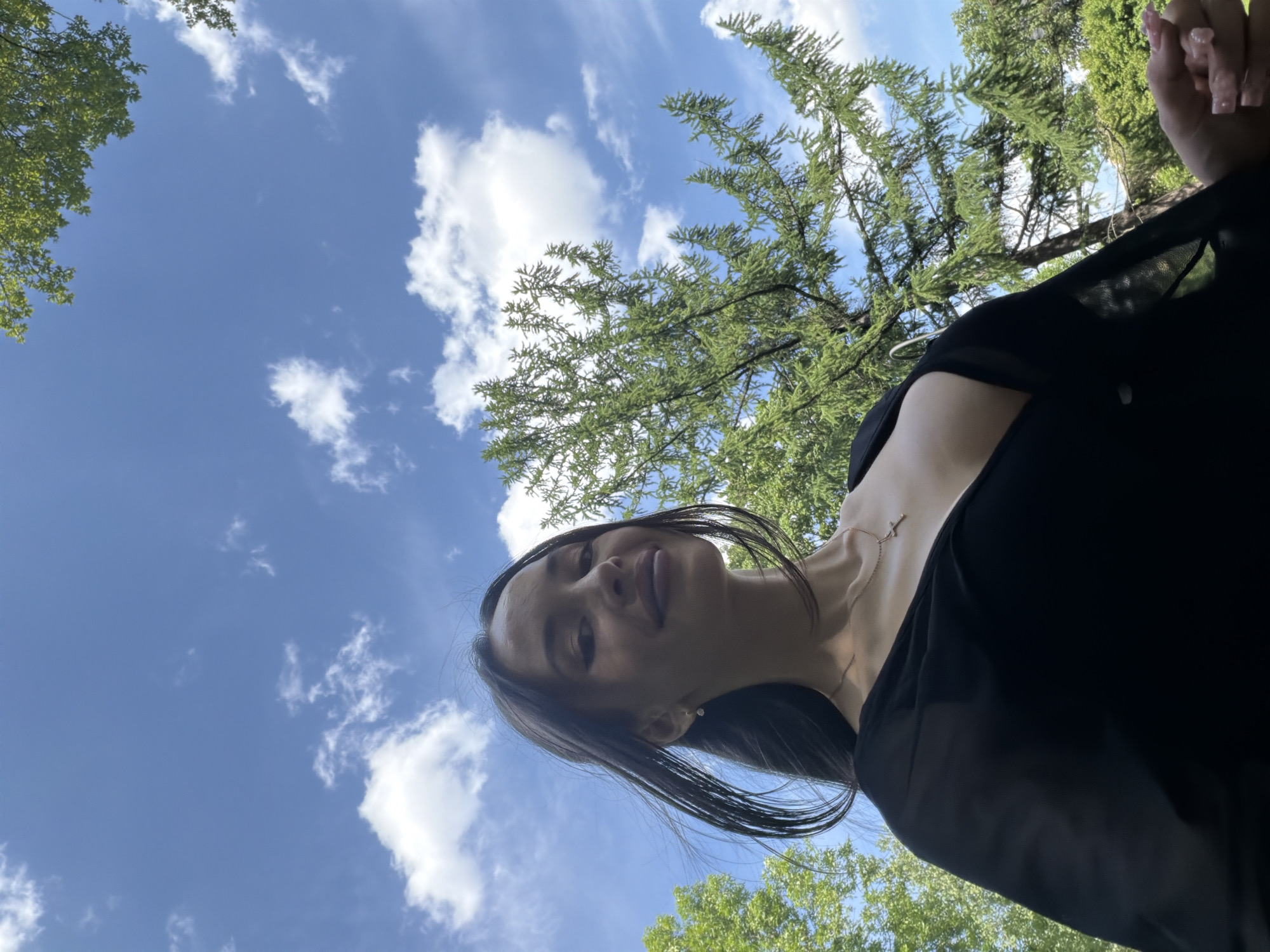 A young adult female is depicted in an outdoor setting, positioned in the lower center of the frame, captured from a low-angle perspective looking upwards. She has long, dark hair, fair skin, and full lips, smiling subtly with her eyes partially closed and dark eyeliner visible. She wears a black top with a deep V-neckline, revealing her upper chest, and a gold-colored chain necklace with a small cross pendant. A small, light-colored stud earring is visible in her right ear. Her right hand, partially visible in the lower right, shows manicured nails with a light, possibly glittery, polish. The background consists of a clear blue sky with scattered white cumulus clouds. Lush green foliage from several trees, including both broad-leafed deciduous trees and needle-leafed coniferous trees, frames the subject and occupies the upper and side portions of the image. The scene takes place in a park or green space in Saint Petersburg, Russia.