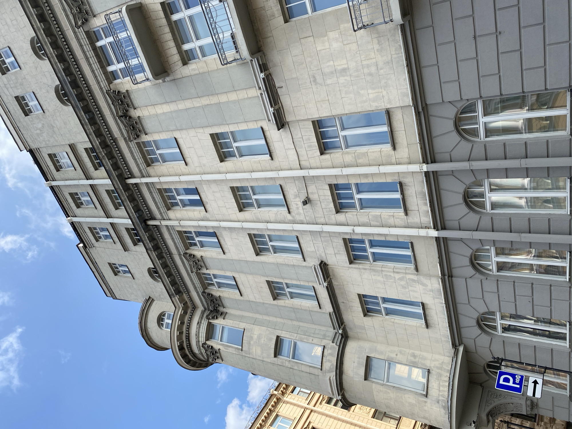 This is a low-angle exterior shot of a multi-story building in Saint Petersburg, Russia. The building has a pale gray, textured facade with numerous windows, many of which are tall and rectangular, while some on the lower floors are arched. The building features classical architectural elements such as cornices and decorative columns. A rounded section with windows is visible on the left side of the building. A blue parking sign with a white "P" and a directional arrow is mounted on a pole in the lower left corner. The sky above is blue with scattered white clouds. The image appears to have been taken during daylight hours.