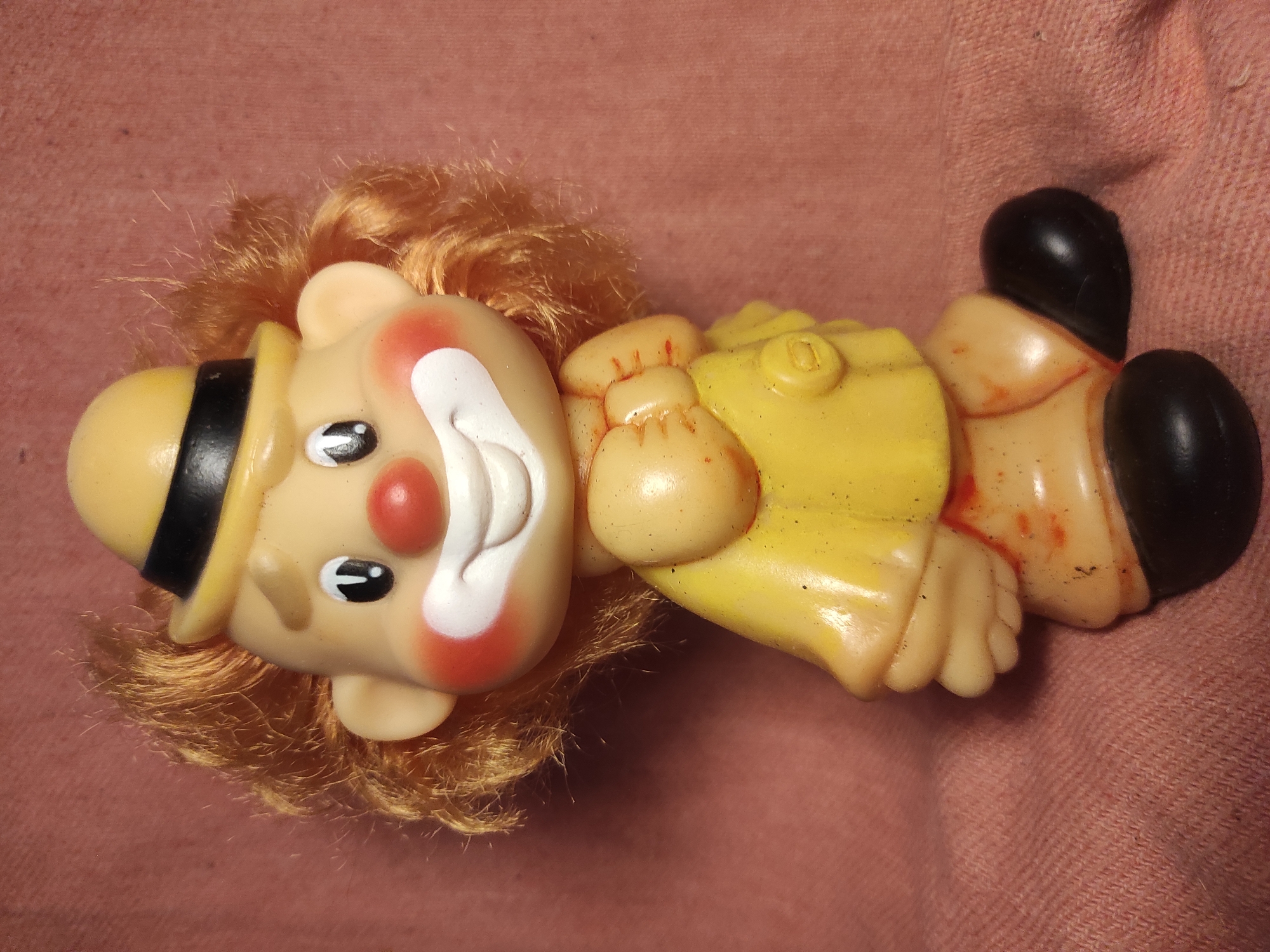 A plastic figurine of a clown with orange hair, a yellow hat, and a yellow suit stands on a pink textured surface. The clown has a smiling face with red cheeks and a red nose. There are smudges of red on the clown's yellow suit and legs. The figurine is positioned in the center of the frame, facing slightly to the left. The background is a plain pink fabric. The image was taken on October 26, 2023, in Chernihiv, Ukraine.