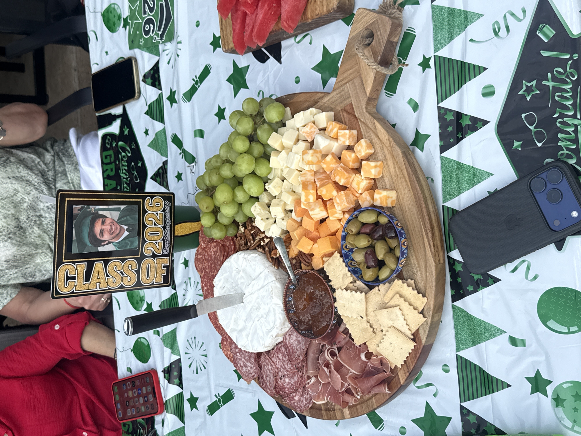 The image shows a wooden charcuterie board laden with a variety of food items, including grapes, cubed cheeses, a wheel of brie with a spoon in an accompanying dish of chutney, olives, crackers, nuts, and various cured meats like salami and prosciutto. The charcuterie board is set on a table covered with a white plastic tablecloth decorated with green graduation-themed designs such as diplomas, stars, and "congrats" messages. A photo frame displaying a graduation photo and the words "Class of 2026" is visible near the board. There are two smartphones on the table, one black and one red, and what appears to be another phone or electronic device in the upper left corner. A hand wearing a red shirt is also visible.
Given the graduation-themed decorations, it is likely that this scene depicts a graduation celebration or party. Without additional context from the image, the location in Southlake, United States of America, cannot be verified.