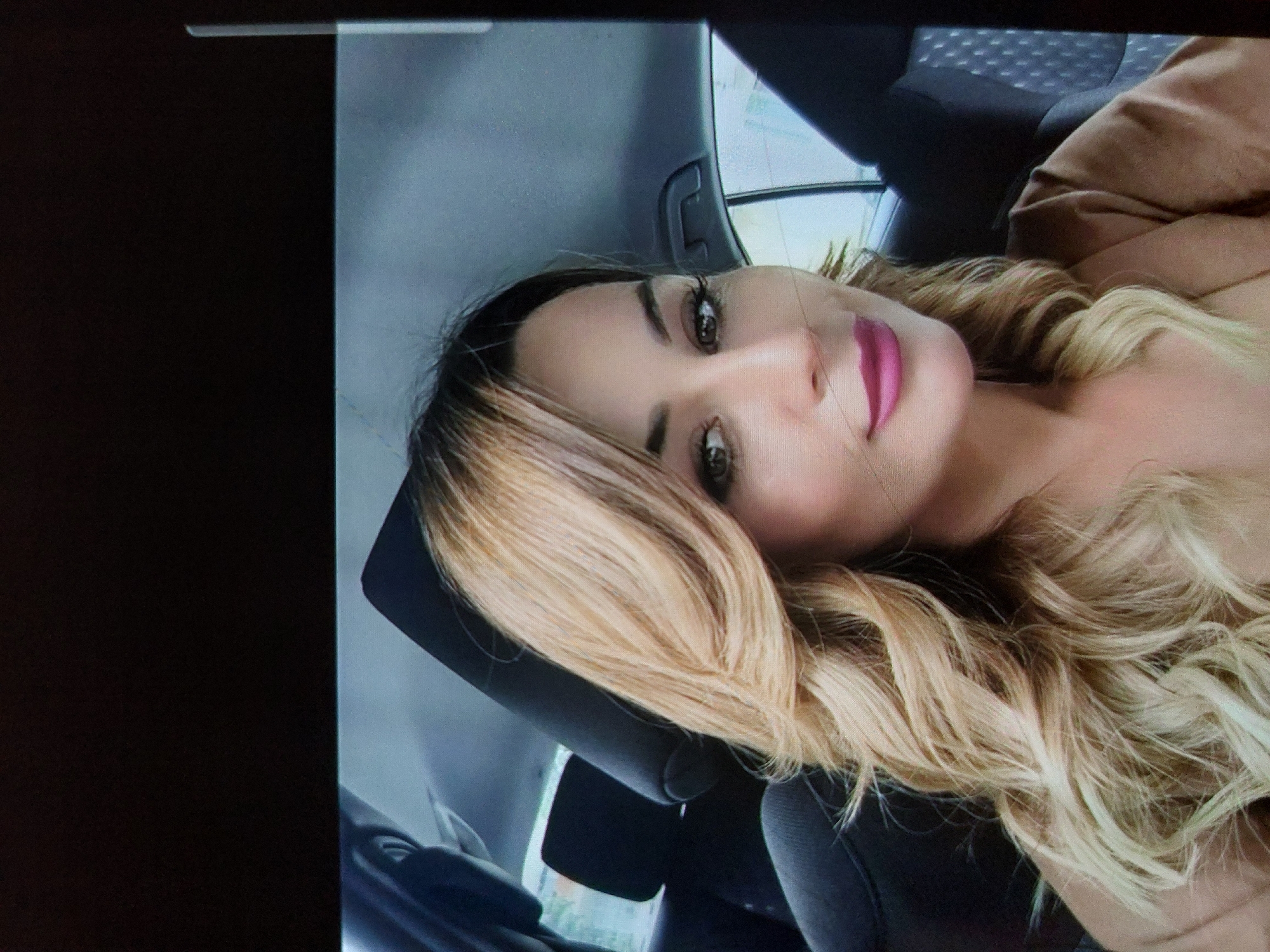 A close-up, portrait-style photograph shows a woman inside a car, likely in Tsarevo, Bulgaria. The woman, depicted from the chest up, has long, wavy blonde hair with darker roots, which cascades over her shoulder. Her head rests against a dark car seat headrest. She looks towards the viewer with green or hazel eyes, accentuated by dark mascara and well-defined eyebrows. Her lips are adorned with bright pink lipstick, forming a gentle smile. She wears a light brown or beige top, possibly off-the-shoulder, visible on her right side. The background includes parts of the car's interior, such as a seat and window frame, with a blurred, sunlit outdoor scene visible through the windows, indicating it is daytime. The photograph appears to be taken of a screen, showing some digital noise and faint horizontal lines. No text is visible in the image.