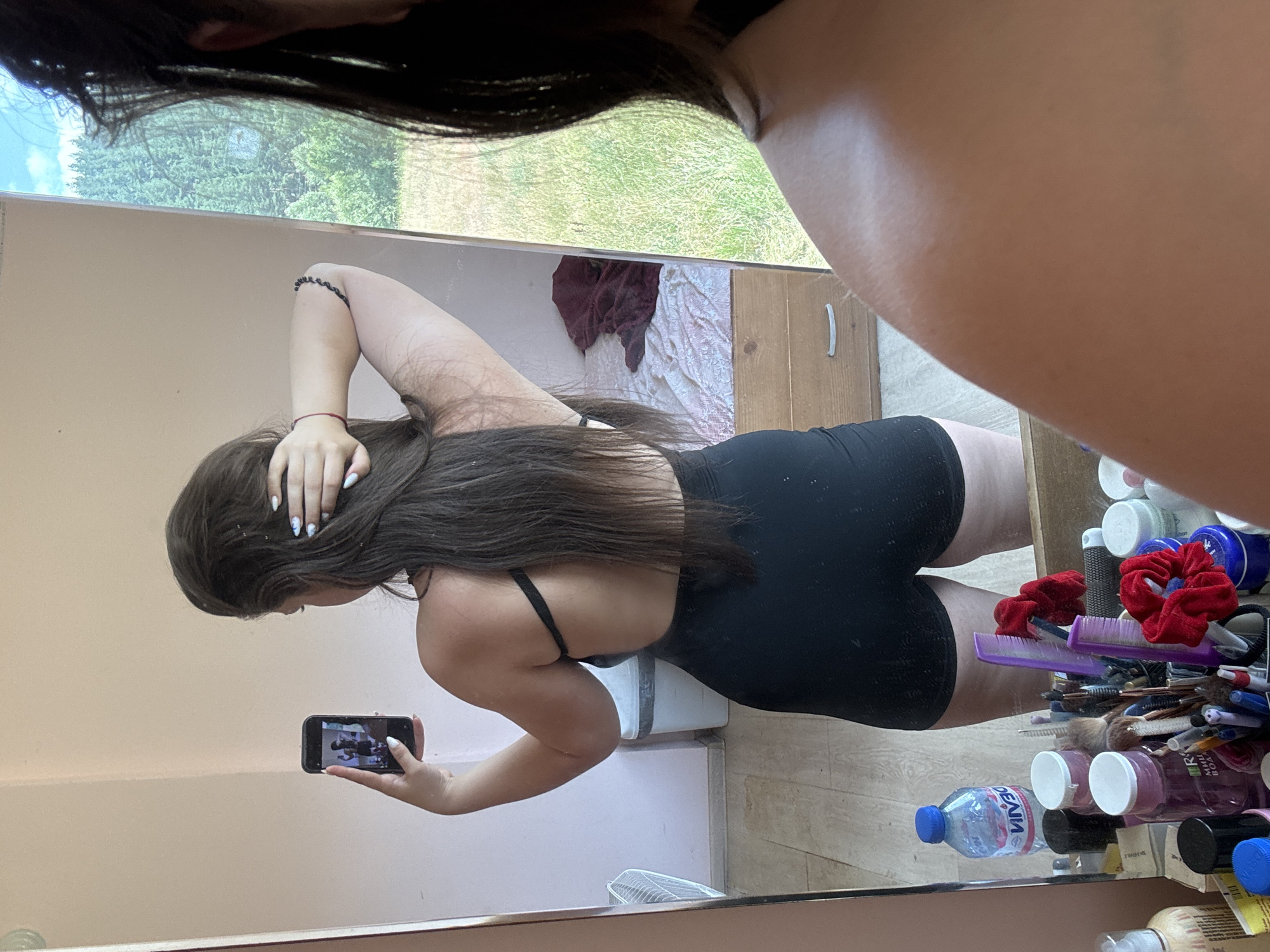 In Prodanovtsi, Bulgaria, a young woman with long, dark hair stands in a room and takes a mirror selfie. She is wearing a black swimsuit or leotard. Her back is to the camera, and she is holding her hair back with one hand, while the other hand holds up a smartphone. The mirror reflects her and part of the room, which includes a bed with a red blanket, a dresser, and a trash can. The background outside the window shows a dense forest under a bright blue sky. In the foreground of the mirror's reflection, a collection of toiletries and hair accessories are visible on a surface.