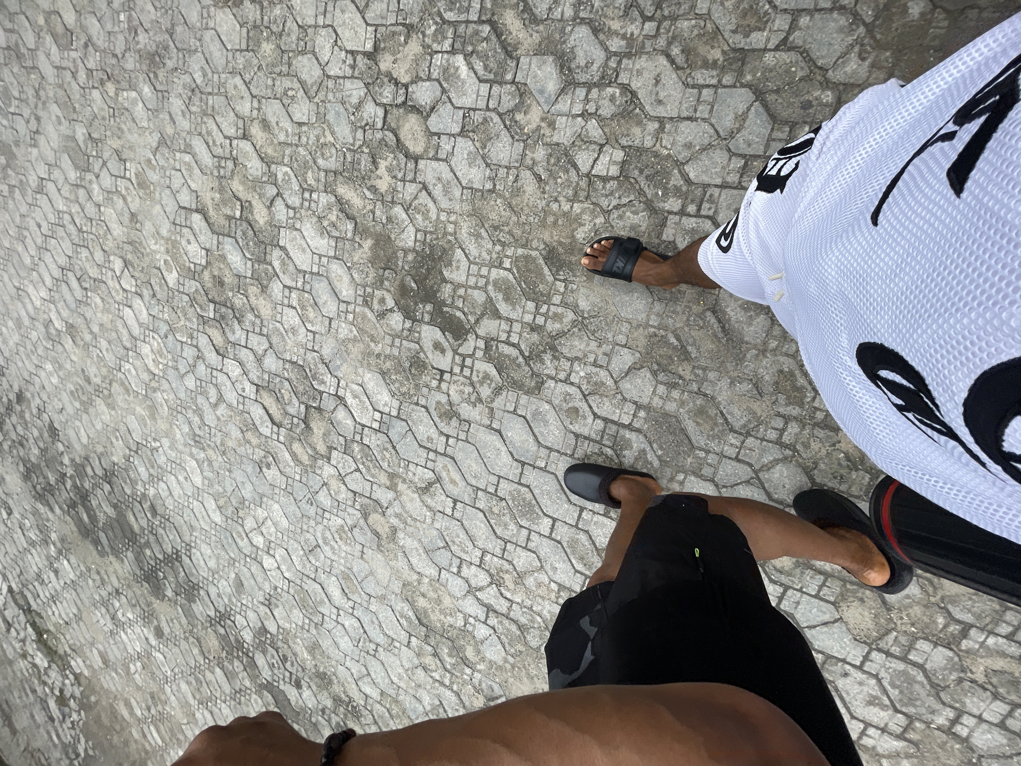 The image shows a first-person, downward view of two people standing on a patterned concrete surface. The surface appears to be outdoors. One person wears a white sports jersey with black graphics and black sandals. The other person wears black shorts and dark sandals. Part of a hand with a bracelet is visible in the foreground. The location is potentially Ado, Nigeria.