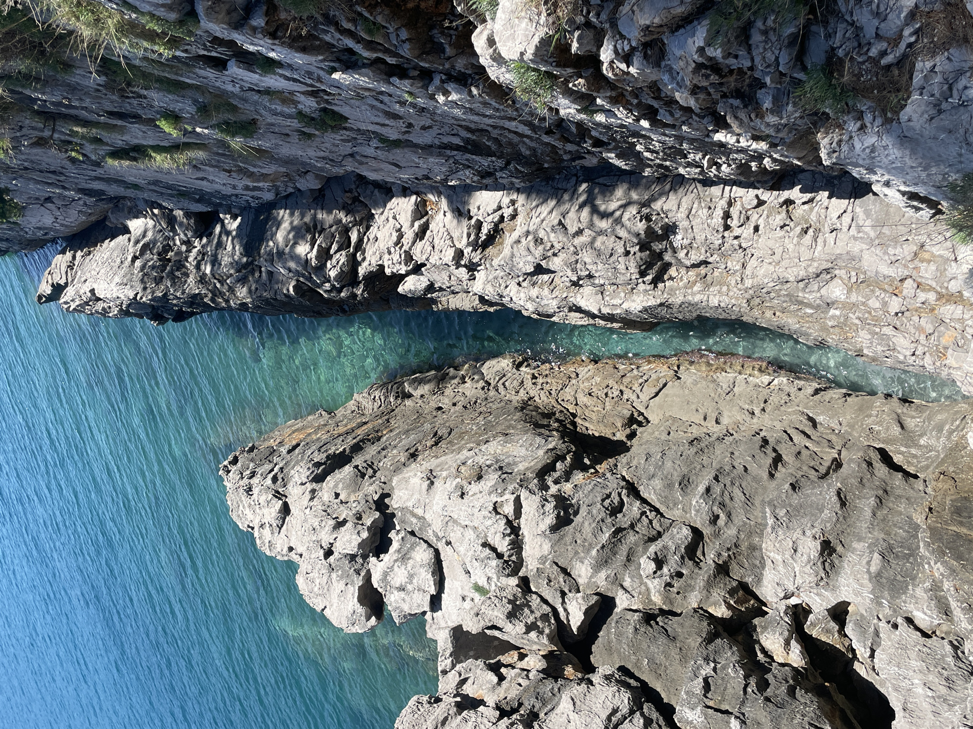 The image presents an aerial or elevated view of a dramatic rocky coastline meeting the sea, likely in Budva, Montenegro. The scene is dominated by rugged, grey rock formations and clear, blue-green water.
A vast expanse of deep blue sea occupies the lower and left portions of the frame, showing gentle ripples on its surface, indicative of calm conditions. Towards the upper right, a narrower, winding inlet or channel cuts into the land, separating two prominent rock masses. The water within this channel is a lighter, crystal-clear turquoise, allowing visibility of the submerged rocky seabed.
The surrounding cliffs are composed of formidable, stratified grey rock, heavily textured with numerous cracks, fissures, and ledges. These formations rise steeply from the water's edge, displaying the raw power of nature. Sparse patches of vibrant green grass and small plants cling tenaciously to the crevices and upper ledges of the rocky slopes, providing small bursts of life against the stone.
The scene is bathed in bright sunlight, casting sharp, distinct shadows on the rock faces, particularly in the crevices and on surfaces oriented away from the light source. This suggests a clear, sunny day, either mid-morning or mid-afternoon. There is no visible human activity or text. The overall impression is one of serene natural beauty and rugged coastal wilderness.