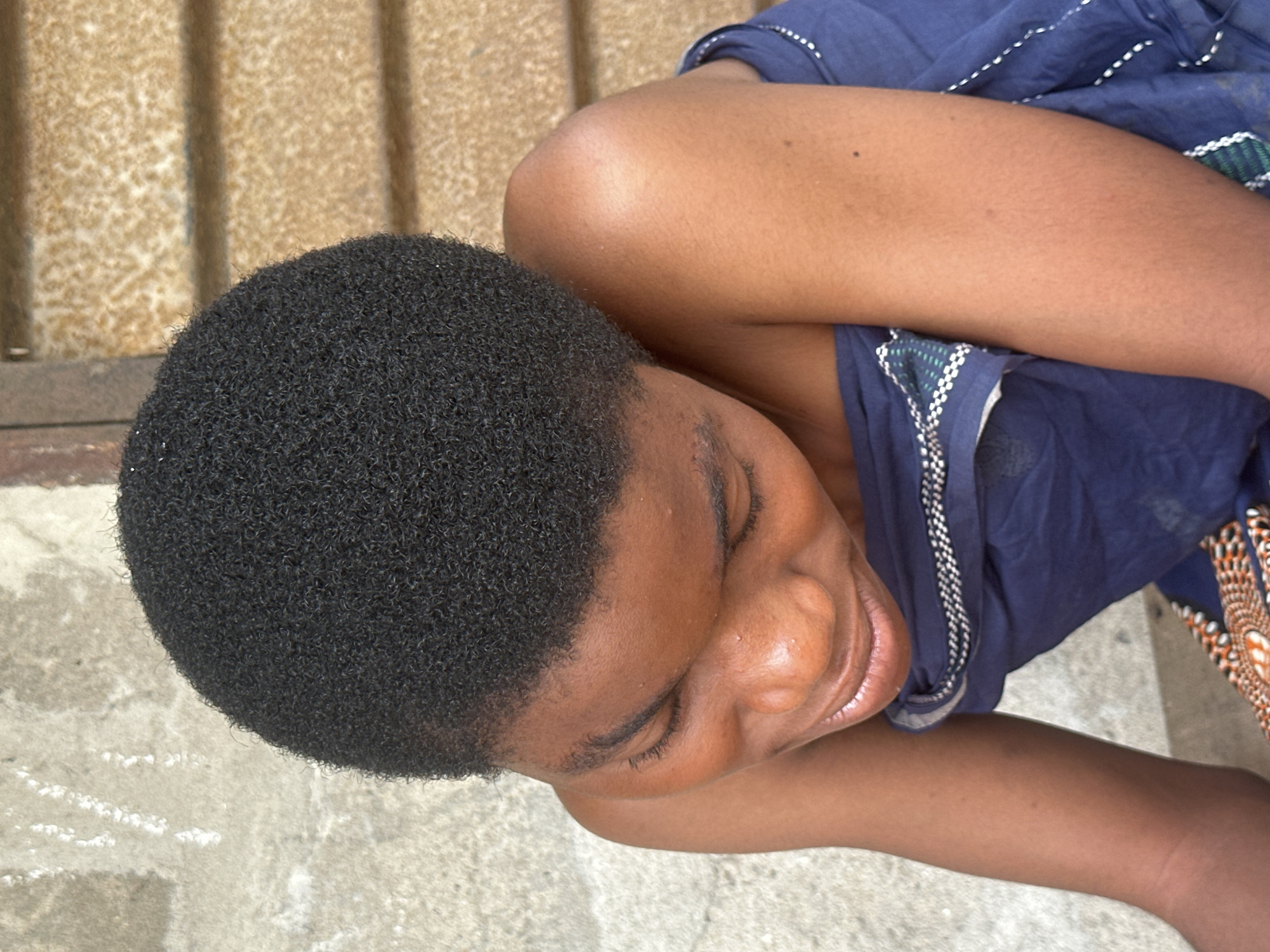 A young Black female, with very short, tightly curled black hair, is captured in an overhead shot in Badagry, Nigeria. She is wearing a dark blue cloth draped around her shoulders and torso, with a patterned trim. Her head is tilted down, and she is looking downwards, with her eyes partially closed and a subtle smile on her lips. Her skin is a warm brown. To the left, a rough, light-colored concrete wall is visible. To the right, horizontal wooden blinds are present. The setting appears to be outdoors, potentially in a residential or public area within Badagry.