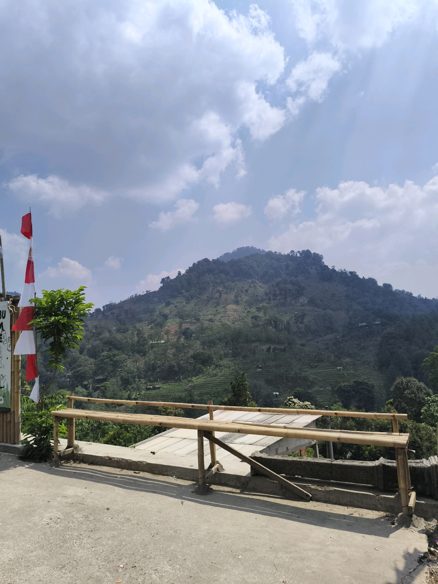 A serene outdoor scene unfolds with a lush, verdant mountainside dominating the background. The mountain is covered in dense foliage, with hints of terraced fields and scattered structures visible on its slopes. A clear blue sky, dotted with fluffy white clouds, stretches above.
In the foreground, a simple bamboo railing runs horizontally across the frame, marking a viewing area. A concrete path or platform is visible beneath the railing, suggesting a constructed overlook. To the left, a small, leafy green tree stands near a vertical bamboo structure adorned with Indonesian flags, adding a touch of local color. The flags, red and white, are partially unfurled, fluttering gently in the breeze.
The overall impression is one of natural beauty and tranquility, perhaps a scenic spot in or near Babakan Madang, Indonesia, offering visitors a picturesque vista of the surrounding landscape.