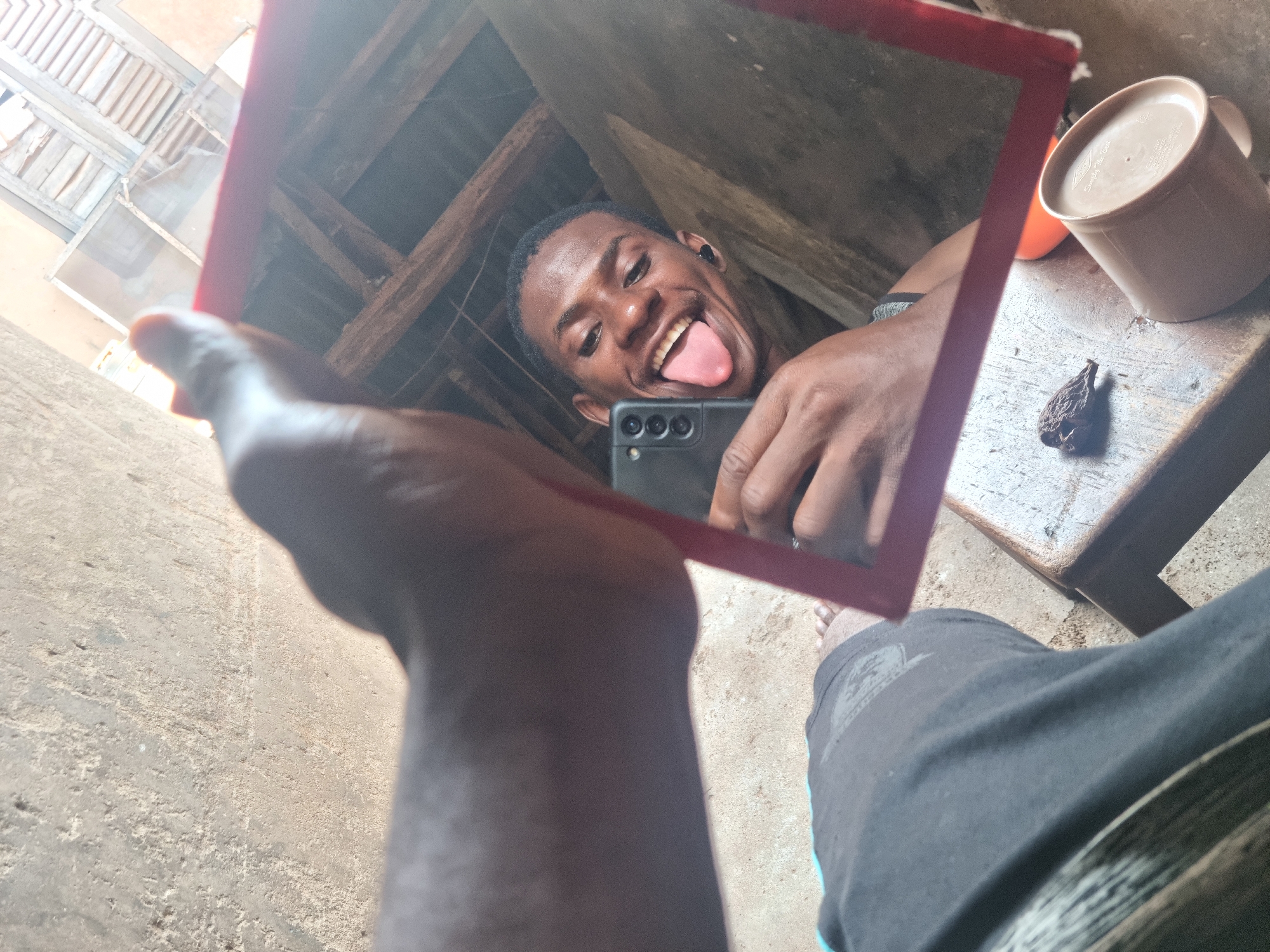 A mirror selfie captures a young man making a playful expression, sticking out his tongue and smiling broadly, reflected within a red-framed mirror. He has short dark hair and is wearing a dark earbud in his right ear. In the reflection, he is holding a black smartphone, likely a Samsung model, visible with its multi-lens camera module.
The background visible through the mirror shows a rustic interior featuring wooden beams and light-colored wooden slats, suggesting a simple roof structure or a part of a building's ceiling/wall. Bright light illuminates this reflected scene, indicating daytime.
Outside the mirror's reflection, a dark-skinned arm and hand hold the red-framed mirror. To the right of the hand, on what appears to be a rustic wooden surface (possibly a stool or small table), rests a light brown mug with faint, illegible text, and a dark, dried, organic object resembling a seed pod or dried fruit. The lower right foreground shows part of a leg wearing dark shorts with a light stripe, and bare feet on a rough, light-colored concrete or dirt floor.
The image conveys a casual, cheerful moment in a modest, sunlit environment, consistent with a setting in Onitsha, Nigeria.
