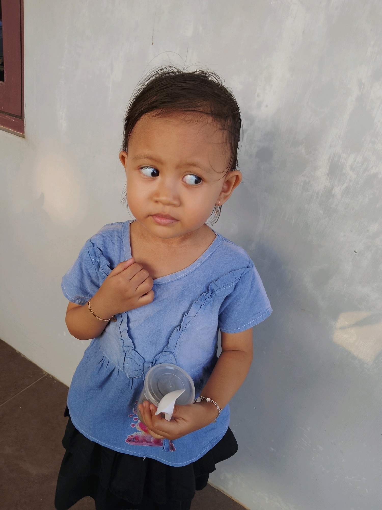 A young child is standing outdoors in front of a textured, light gray wall. The child is wearing a light blue, short-sleeved top with ruffled details and a black skirt. The child is holding a clear plastic container with a white object protruding from it in their right hand. The child's left hand is touching their chest. A gold-colored bracelet is visible on the child's left wrist. The child's head is turned to the left, looking away from the camera with a thoughtful expression. Part of a dark brown window frame is visible on the far left of the image. The ground in the foreground is made of brown tiles. The image appears to have been taken in Pelaihari, Indonesia.