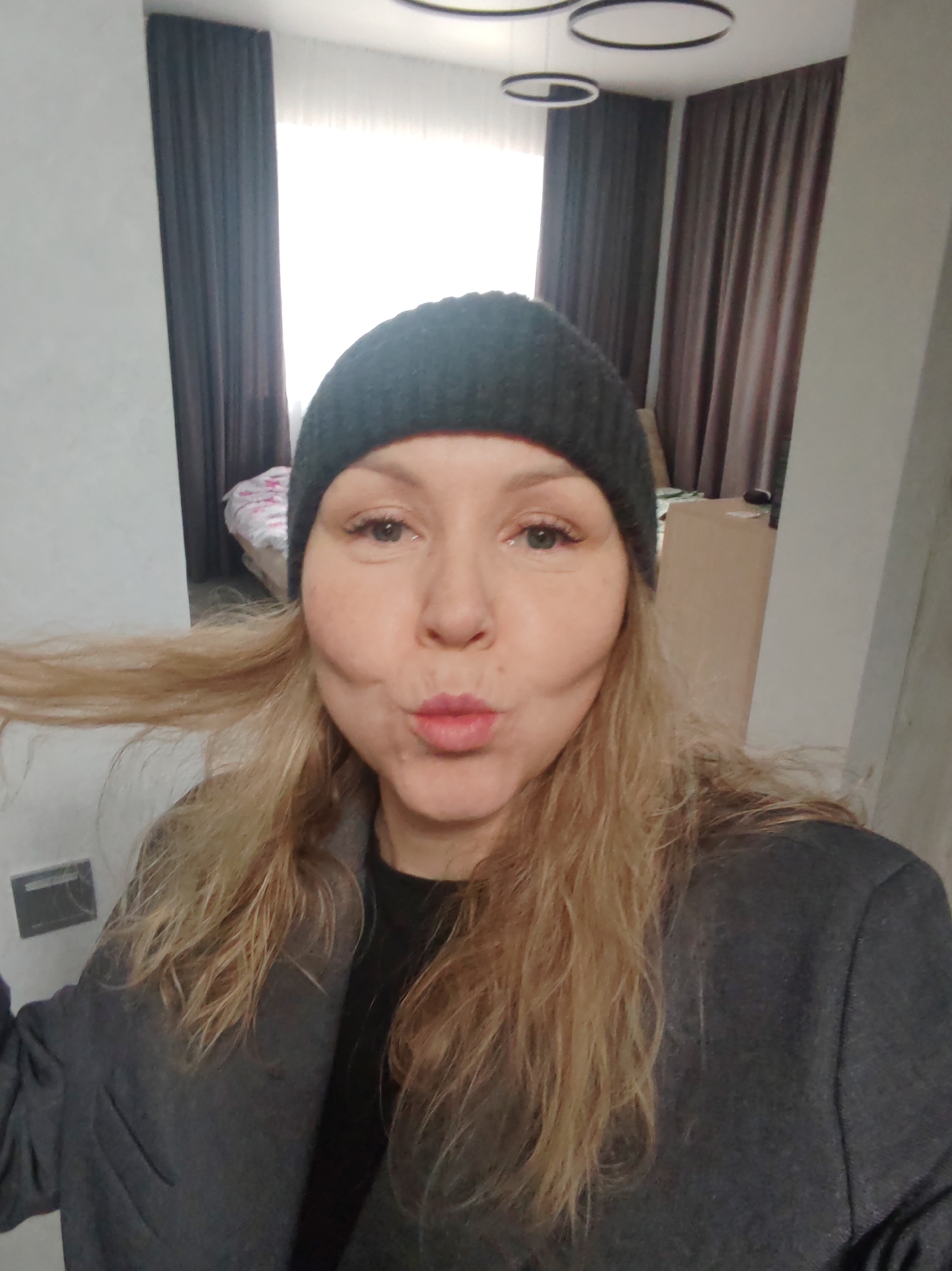 A medium, eye-level shot shows a blonde woman indoors wearing a dark grey beanie and a grey jacket. She is posing with pursed lips. Background elements include a bed with pink and white linens, dark brown curtains, and a modern circular light fixture. The woman appears to be in a domestic setting, possibly an apartment or home. The exact location in Chaikovsky, Russia cannot be determined from the image.