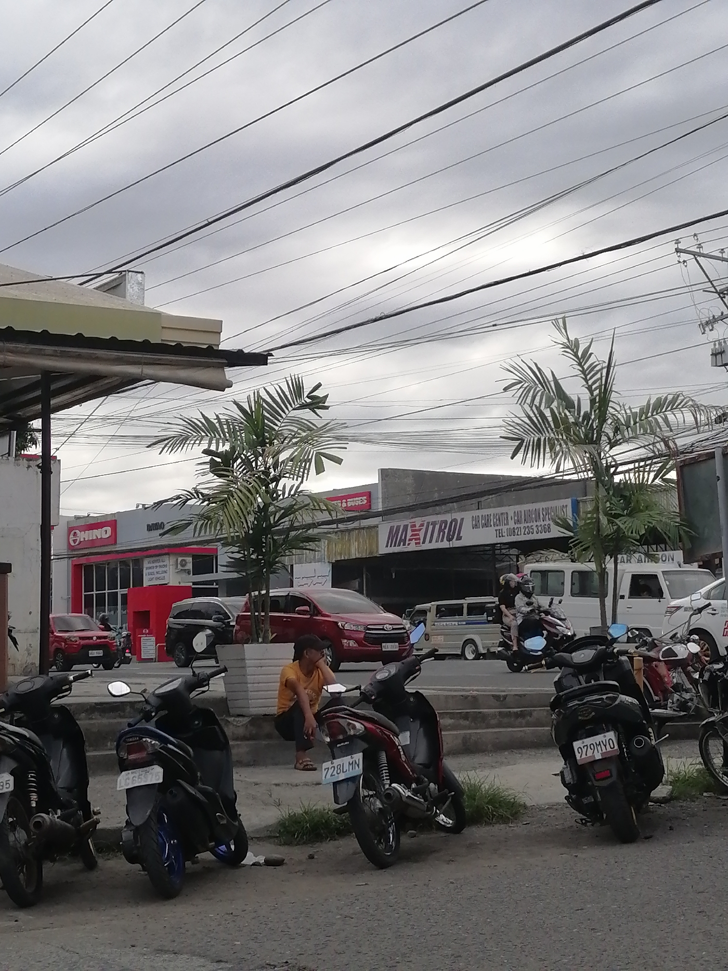 In Davao City, Philippines, a street scene unfolds under an overcast sky, with numerous overhead power lines crisscrossing the frame. On the left, a red Hino dealership is visible, with a red SUV parked outside. Adjacent to it is a building with signage reading "MAXITROL CAR CARE CENTER - CAR AIRCON SPECIALIST." Several parked motorcycles are prominent in the foreground, some with visible license plates. A person in an orange shirt is seated on a ledge near the motorcycles. In the background, a white van and other vehicles are present on the road. The overall environment suggests a commercial area with automotive services.