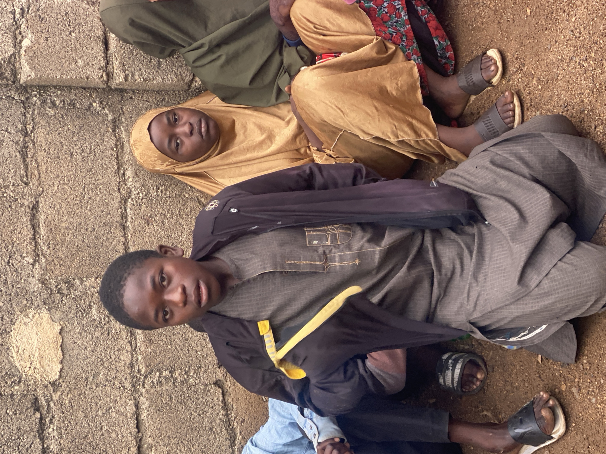 The image shows a group of people, likely children, sitting in front of a rough, concrete wall. The wall appears weathered and textured, with some areas showing exposed aggregate. The ground is covered in sandy soil and small pebbles. In the foreground, a young person wearing a dark jacket over a traditional gray outfit is looking towards the camera. The jacket has a bright yellow object attached to the left sleeve, possibly a piece of fabric or a tool. To the right of this person, another individual is wearing a gold-colored hijab and a long, flowing garment. Further to the right, a third person is visible wearing a dark green hijab and a similar long garment. The clothing suggests a cultural context, possibly related to the attire worn in parts of Nigeria. The overall lighting suggests an outdoor setting, possibly during the daytime. The image captures a candid moment of the individuals seated together.