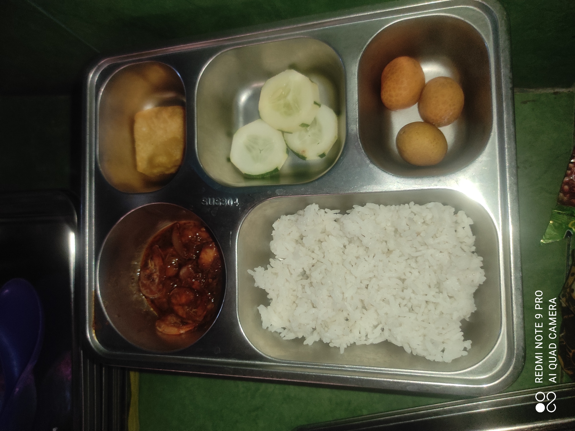 A stainless steel compartmentalized meal tray holds a meal. The top left compartment contains shrimp in a dark sauce. The top right compartment has a piece of fried tofu. The middle right compartment holds three slices of cucumber. The bottom right compartment contains three small, oval-shaped fruits, possibly longans. The large compartment on the left is filled with white rice. The tray is placed on a green surface, and a partial view of a dark, possibly purple, utensil is visible at the top of the frame. The image was taken in Sakra, Indonesia.