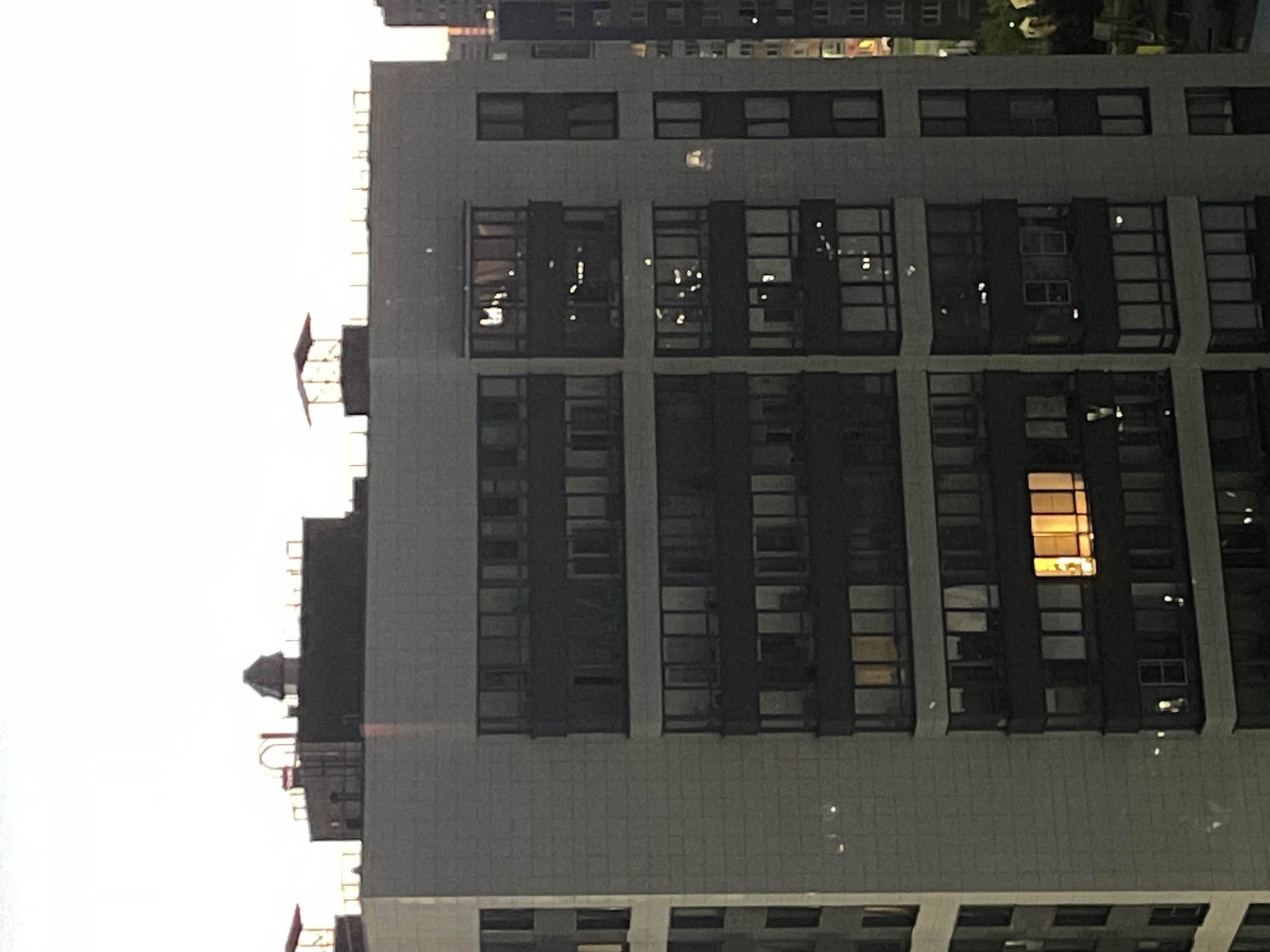 The image captures a close-up, aerial view of a modern apartment building in Sinyavino, Russia. The building features a light-colored facade with dark-framed windows arranged in vertical columns and horizontal rows. Several windows are illuminated from within, suggesting occupancy, with one particularly bright orange light emanating from a window on the right side of the central section. The top of the building reveals elements of the roof structure, including ventilation systems and decorative caps, with a distinctly peaked, dark structure at the highest point of the visible roofline. In the distance, other buildings, also appearing to be apartment blocks, can be seen, some also showing illuminated windows. The sky above is a pale, overcast hue, indicative of twilight or an overcast day. The overall impression is one of urban residential architecture.