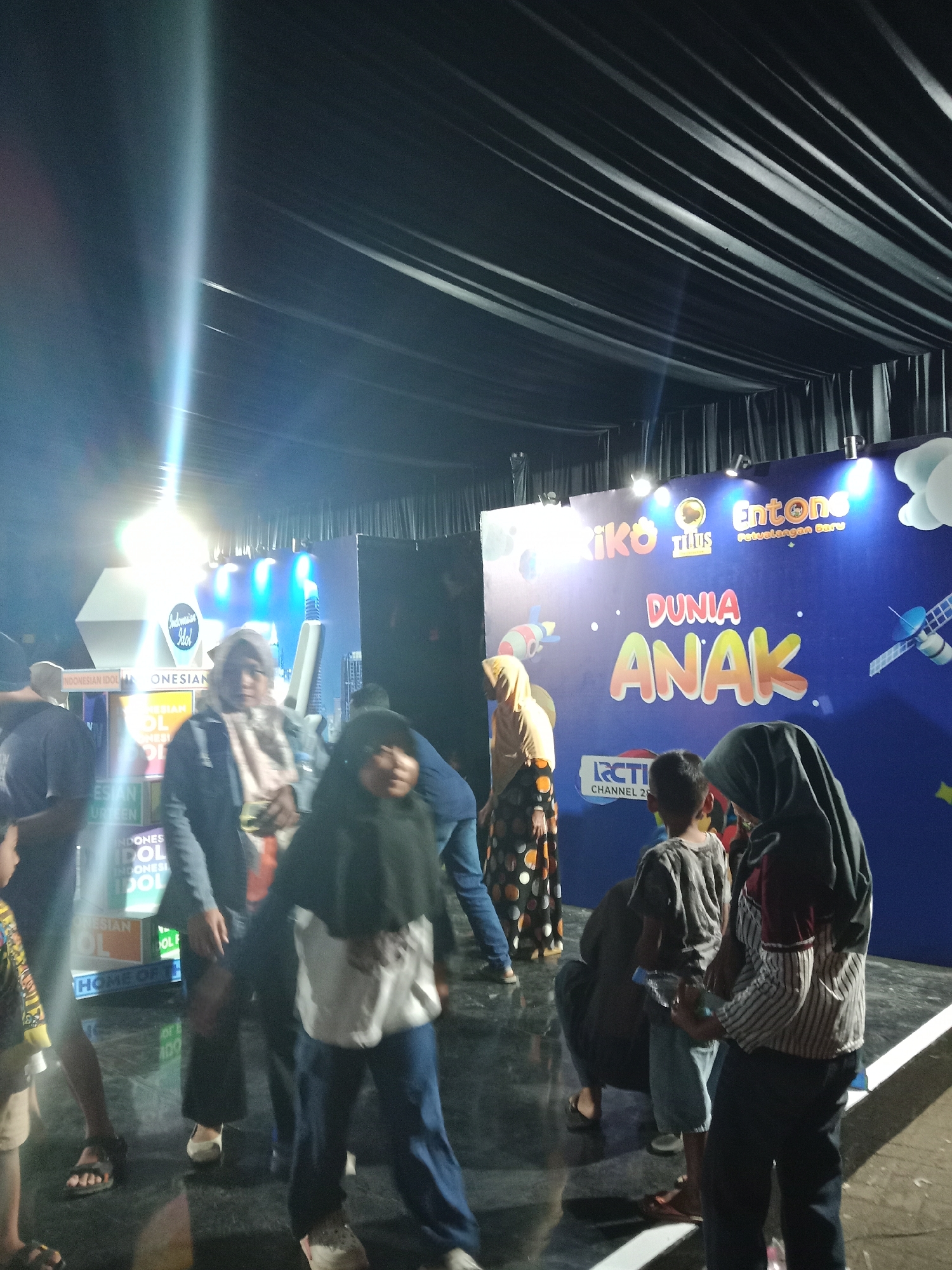 The image depicts an indoor event, possibly a stage or exhibition area, with a large blue backdrop featuring animated characters and text. The backdrop displays the words "DUNIA ANAK" (Children's World) in large, colorful letters, along with logos for "Entong Petualangan Baru" and "RCTI CHANNEL 27". The lighting suggests it is nighttime. Several people are visible, some in traditional Indonesian attire like hijabs, interacting in the foreground and midground. To the left of the backdrop, there's a structure with "INDONESIAN IDOL" branding. The environment appears to be a public gathering space, possibly a convention center or a similar venue in Kendal, Indonesia, where this event is taking place.