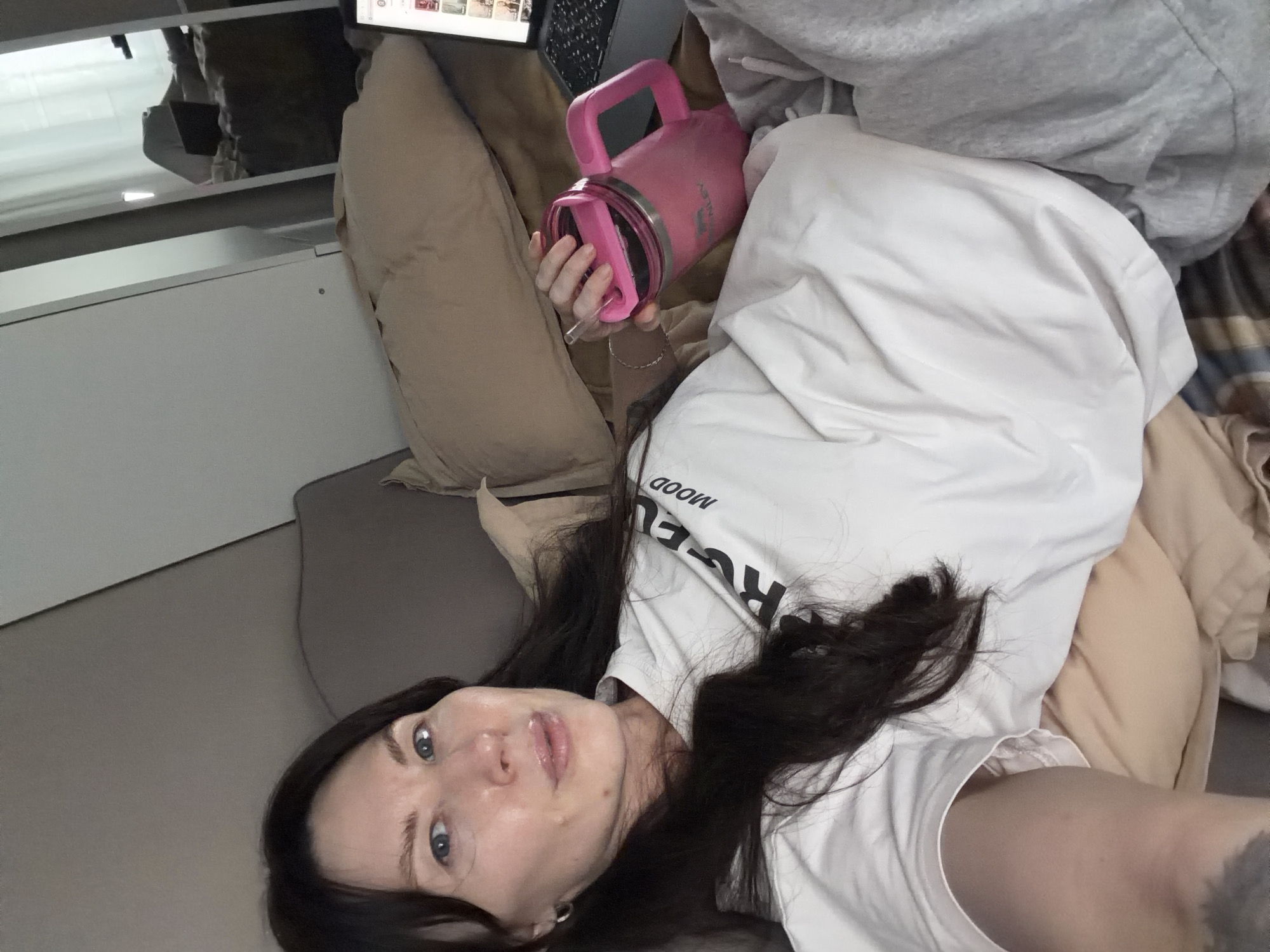 A female individual is depicted in a close-up, elevated selfie taken indoors, possibly in a bedroom or lounge area. She is positioned lying down or reclining against tan-colored pillows and bedding, with a grey upholstered headboard or cushion visible behind her. The subject has dark brown hair, blue eyes, and fair skin, appearing to have minimal to no makeup, with a slight sheen on her lips. She wears a white t-shirt with dark lettering, part of which reads "MOOD," and light grey sweatpants. A dark tattoo is partially visible on her left upper arm. Her right hand grasps a pink Stanley-brand insulated mug with a handle and a pink straw. A laptop is open on her right side, displaying a screen with what appears to be a web page featuring multiple small images or video thumbnails. The background includes a white wall or door segment and a reflective surface, possibly a mirror, showing a light-colored curtain or window arrangement. No specific landmarks or identifying features are present to determine the precise location within Perm, Russia.