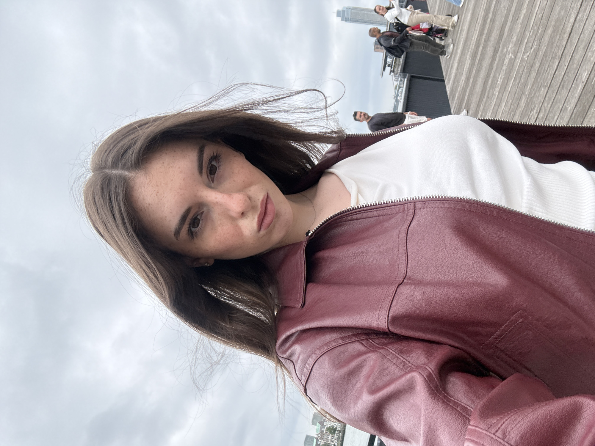 A woman with long brown hair and freckles, wearing a burgundy leather jacket and a white top, takes a selfie on a cloudy day. In the background, people walk along a wooden pier with a city skyline visible across the water. The architecture suggests the location is likely in a harbor area of a major city. The view is consistent with areas like Kop van Zuid in Rotterdam, Netherlands, known for its modern buildings and waterfront promenades.