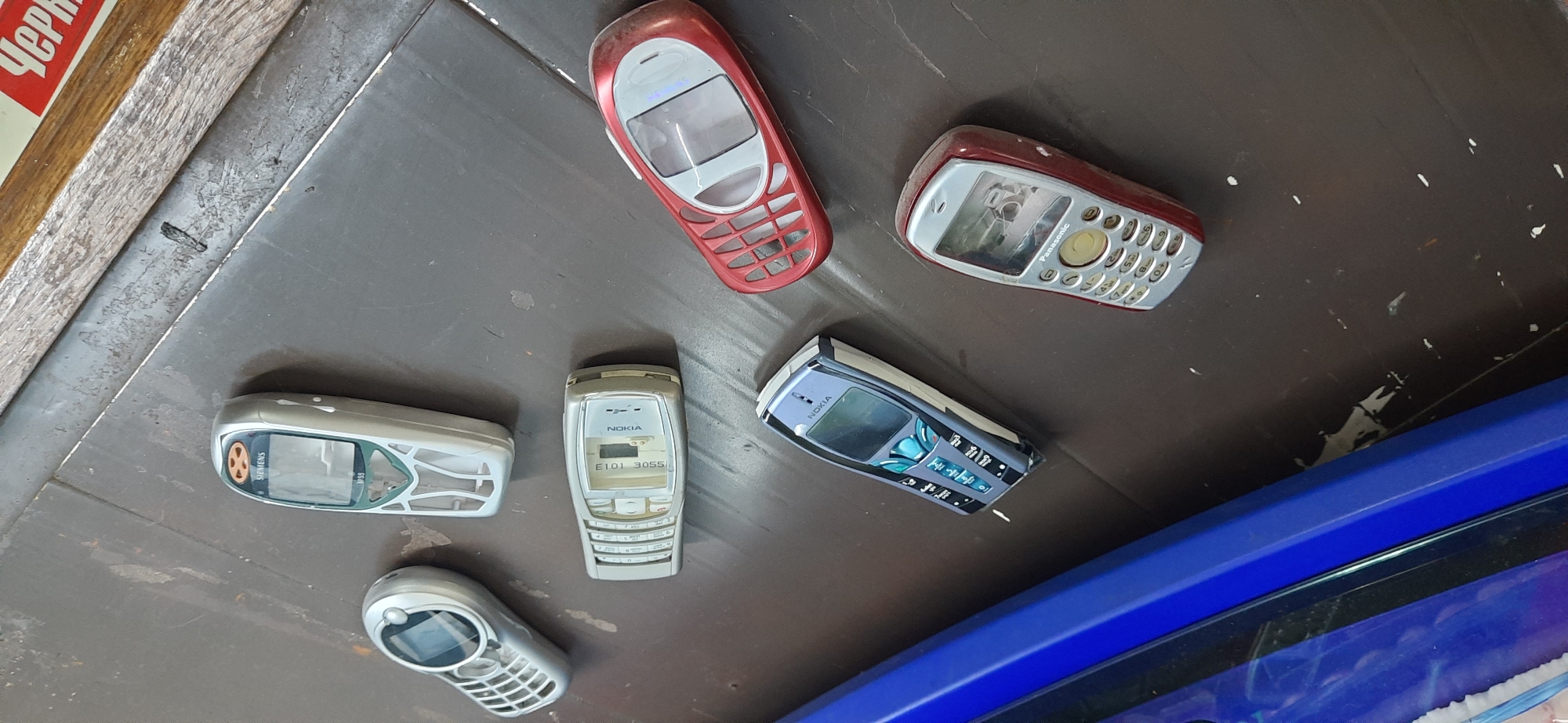 The image displays a collection of seven old mobile phones scattered across a dark, textured surface, possibly a table or floor. The phones are of various brands, including Siemens, Nokia, and Panasonic. The Siemens phone, in the upper left, is silver and white with a clear screen and a silver keypad. Next to it, a silver and white Nokia phone is lying flat, showing a green keypad and a screen. Below this, another Nokia phone, elongated and silver, has "E101 3055" visible on its screen. To the right, a red and silver phone, likely a Nokia, is positioned diagonally, featuring a rounded shape and an open grille design. Further down, a blue and silver Nokia phone with a distinctly futuristic design is visible. Another silver Panasonic phone, with a circular navigation button and a numeric keypad, is situated in the lower right. Finally, a red and silver phone with a similar keypad layout to the Panasonic is to the left of it. The background shows a wooden surface with some light-colored debris. The scene does not provide any specific landmarks or elements that would definitively place it within the city of Dnipro, Ukraine. The lighting suggests an indoor environment, likely with ambient light.