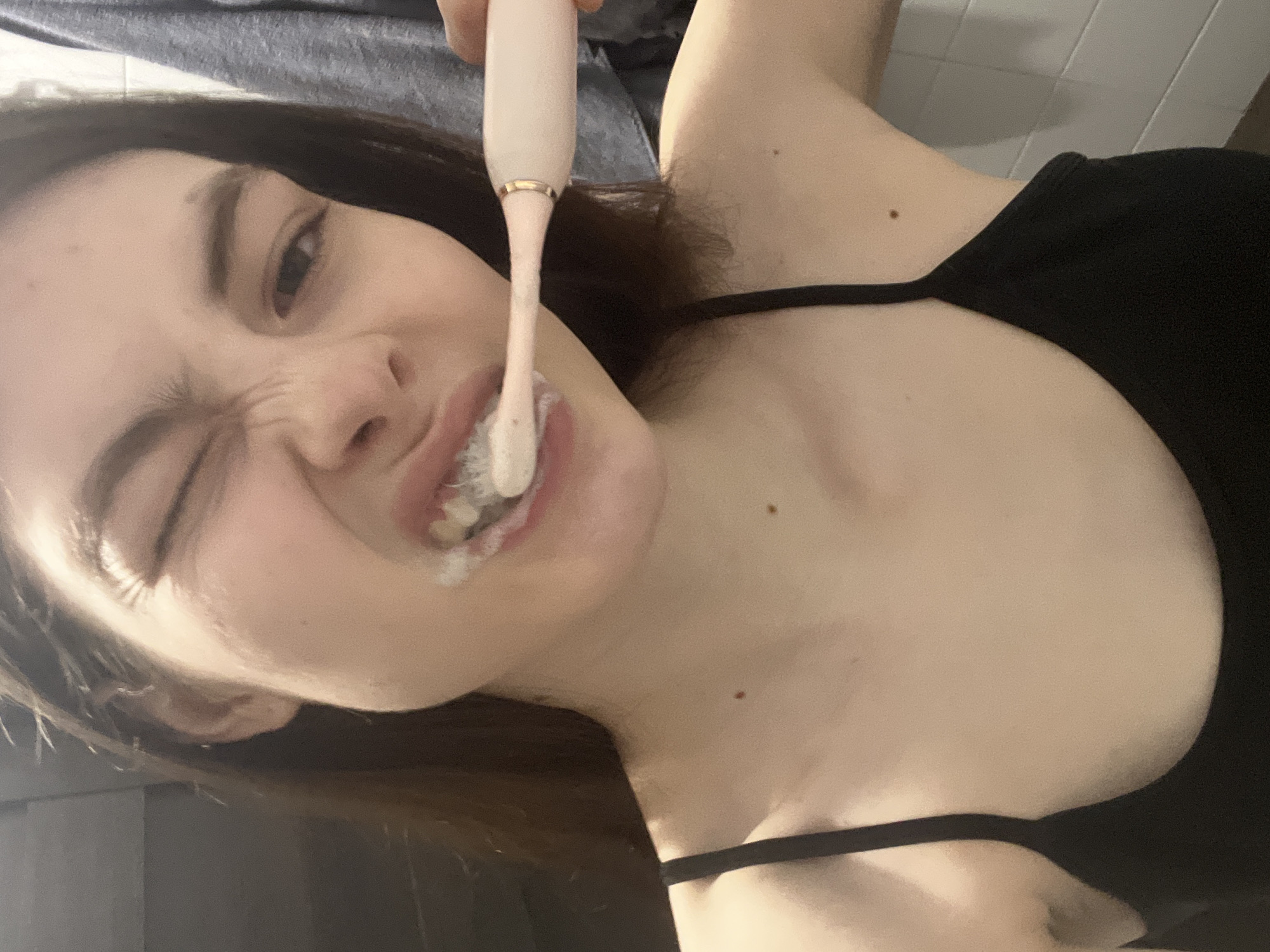 A woman is brushing her teeth with an electric toothbrush. She is wearing a black tank top. The background is blurry, showing what appears to be a bathroom with light-colored tiles. There is no evidence to indicate a location in the city of Saint Petersburg, Russia.