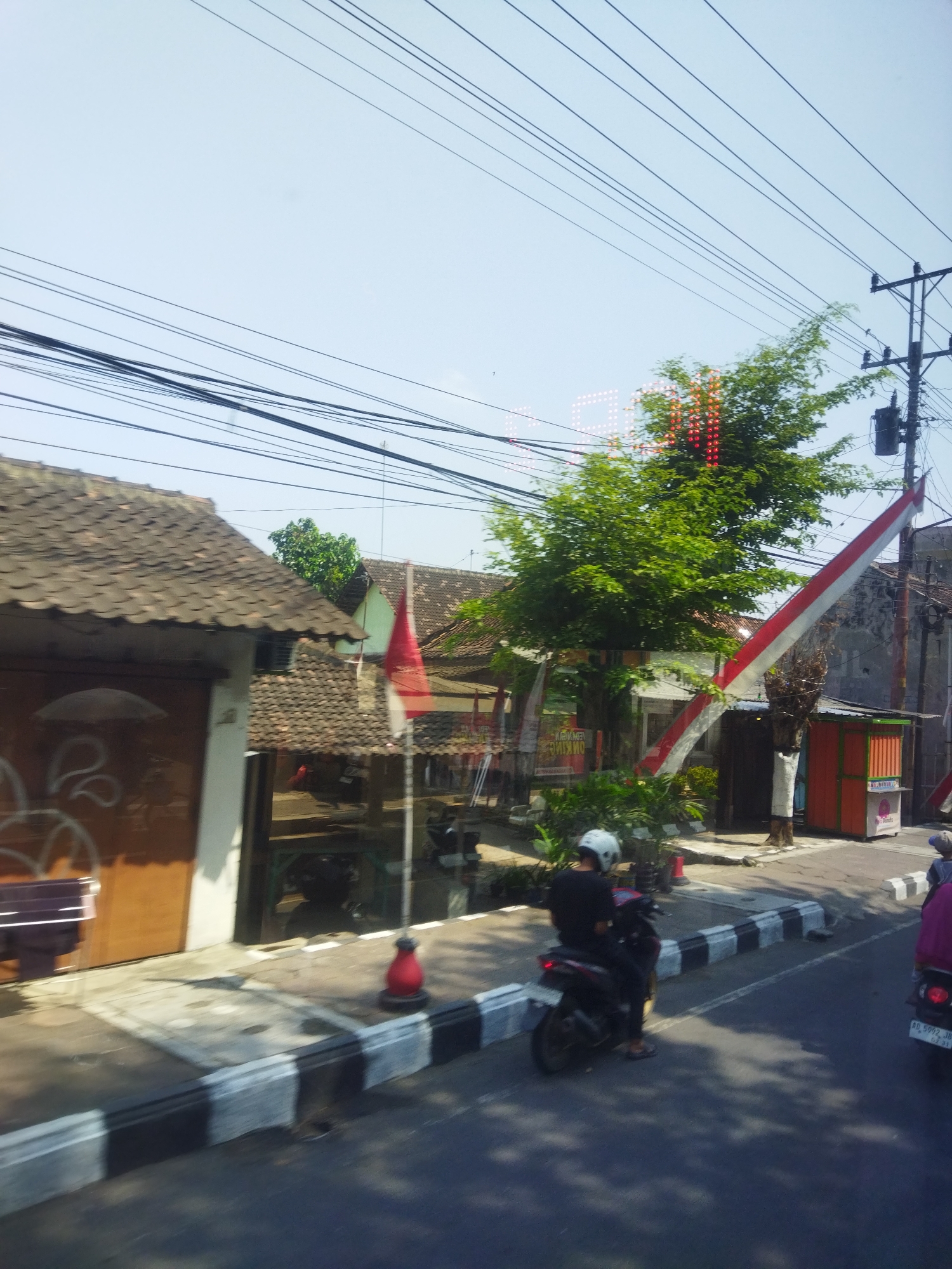 This is a street-level, outdoor, medium shot of a street in Jombor, Indonesia, likely taken from a moving vehicle. The sky is bright and clear, with numerous power lines crisscrossing overhead. On the left side of the street, there are several buildings with tiled roofs. One building has a large, brown-colored door with some white graffiti. Adjacent to this building is a smaller structure with a reddish-brown tiled roof. Further down the street, a red and white Indonesian flag is visible, attached to a tree.
In the foreground, a person wearing a black shirt and white helmet is riding a black motorcycle. The motorcycle is parked on the side of the road, next to a curb with black and white stripes. A red traffic cone is visible near the curb. To the right of the motorcycle rider, another motorcycle, partially visible, is being ridden by a person wearing a blue shirt and a grey hat. This motorcycle has a license plate that reads "AG 5092 JB".
The street is paved and appears to be in moderate condition. Sunlight casts shadows on the road and sidewalk, indicating it is daytime. There is greenery in the background, suggesting a residential or commercial area with trees and plants. The overall impression is a typical Indonesian street scene on a sunny day, possibly during a national holiday due to the presence of the flag.