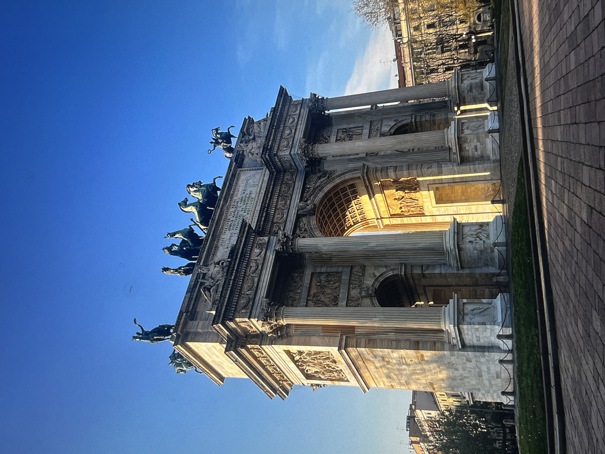 Here is a description of the image:
The image shows the Arco della Pace (Arch of Peace) in Milan, Italy. The arch is a neoclassical triumphal arch adorned with sculptures and inscriptions. The bronze sculpture of the Sestiga della Pace (the Peace Sestiga) is on top of the arch. The arch is made of marble. The sky is blue.