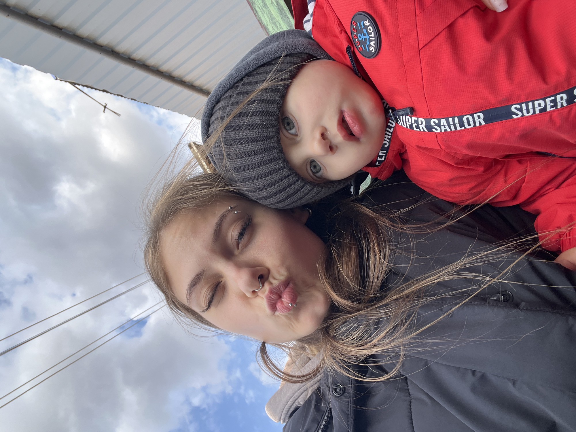 A woman and a young child are posing for a selfie. The woman has light skin, brown hair, and facial piercings. She is puckering her lips towards the camera. The child is wearing a gray beanie and a red jacket with "SUPER SAILOR" printed on it. The child is looking at the camera with its mouth open and tongue sticking out slightly. The background shows a cloudy sky and what appears to be a metallic or corrugated roof. The location is not discernible as Фоки, Russia.