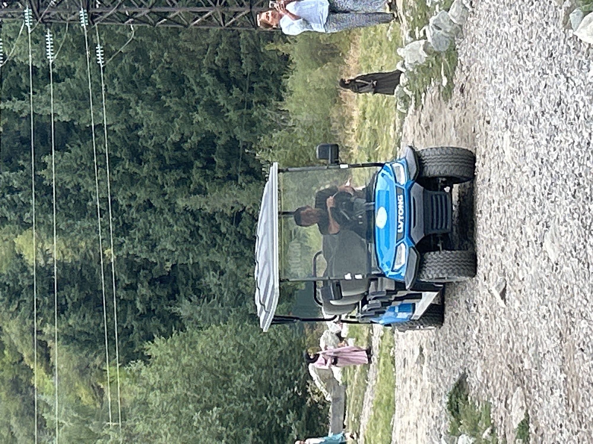 The image captures a sunny day scene on a gravel path in the mountains near Almaty, Kazakhstan. In the foreground, a blue golf cart is parked, driven by a man. A woman in a light blue shirt and patterned pants stands to the right, holding a smartphone and taking a picture. Further in the background, a woman in a pink headscarf and dress walks away from the camera, and another woman in a dark veil stands near a pile of rocks. The path is surrounded by lush green trees and vegetation, with power lines visible overhead. The overall impression is of a tourist or recreational setting within a natural landscape.