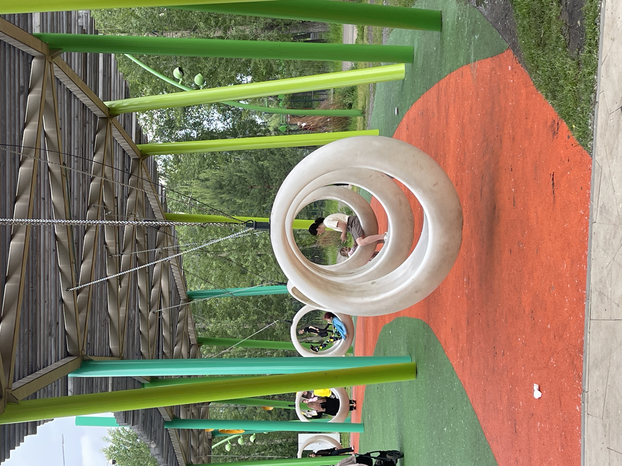 A brightly colored children's playground in the city of Krasnoyarsk, Russia, features a unique circular swing set. The swings are large, white, ring-shaped structures suspended from chains, offering a contemporary aesthetic. The playground surface is a vibrant mix of green and orange rubberized material, with a curved orange path guiding visitors through the area. Tall, slender green and lime-colored posts support a slatted, grey roof structure, creating a shaded pavilion over the swings. In the background, lush green trees provide a natural backdrop. Several people are visible interacting with the playground: a woman sits on one of the swings, holding a child; another person is seated in a different swing, while others stand nearby, observing or waiting. A stroller is parked on the green surface to the left of the frame. The scene captures a lively, public space designed for recreation.
