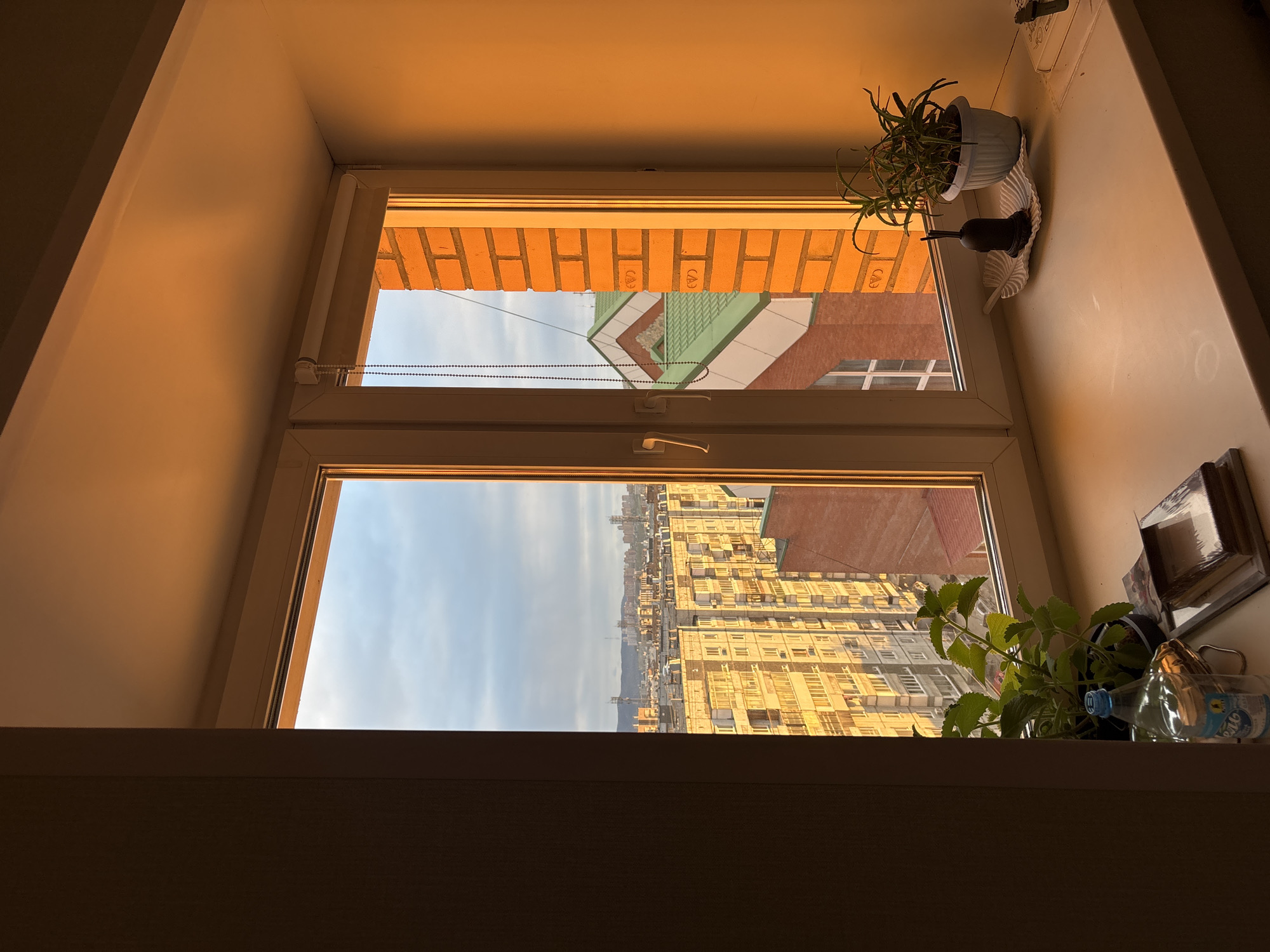 The image shows a window in what appears to be a room, overlooking a cityscape. The window has a white frame, and is divided into two panes. The view through the top pane shows a close building with brick and a green roof. The lower pane gives a wider view of the city with dense buildings in yellow hues, possibly reflecting sunlight. A blue sky is visible above the buildings. On the windowsill, to the right, there are two plants, one in a white pot and the other in a black pot, as well as some decorative objects and a water bottle. The room is lit by warm light, and the overall impression is one of a cozy interior space looking out onto a bustling urban landscape. Based on visual analysis, the city appears to match the architecture of Krasnoyarsk, Russia, with its distinctive buildings. No people are visible in the image.
