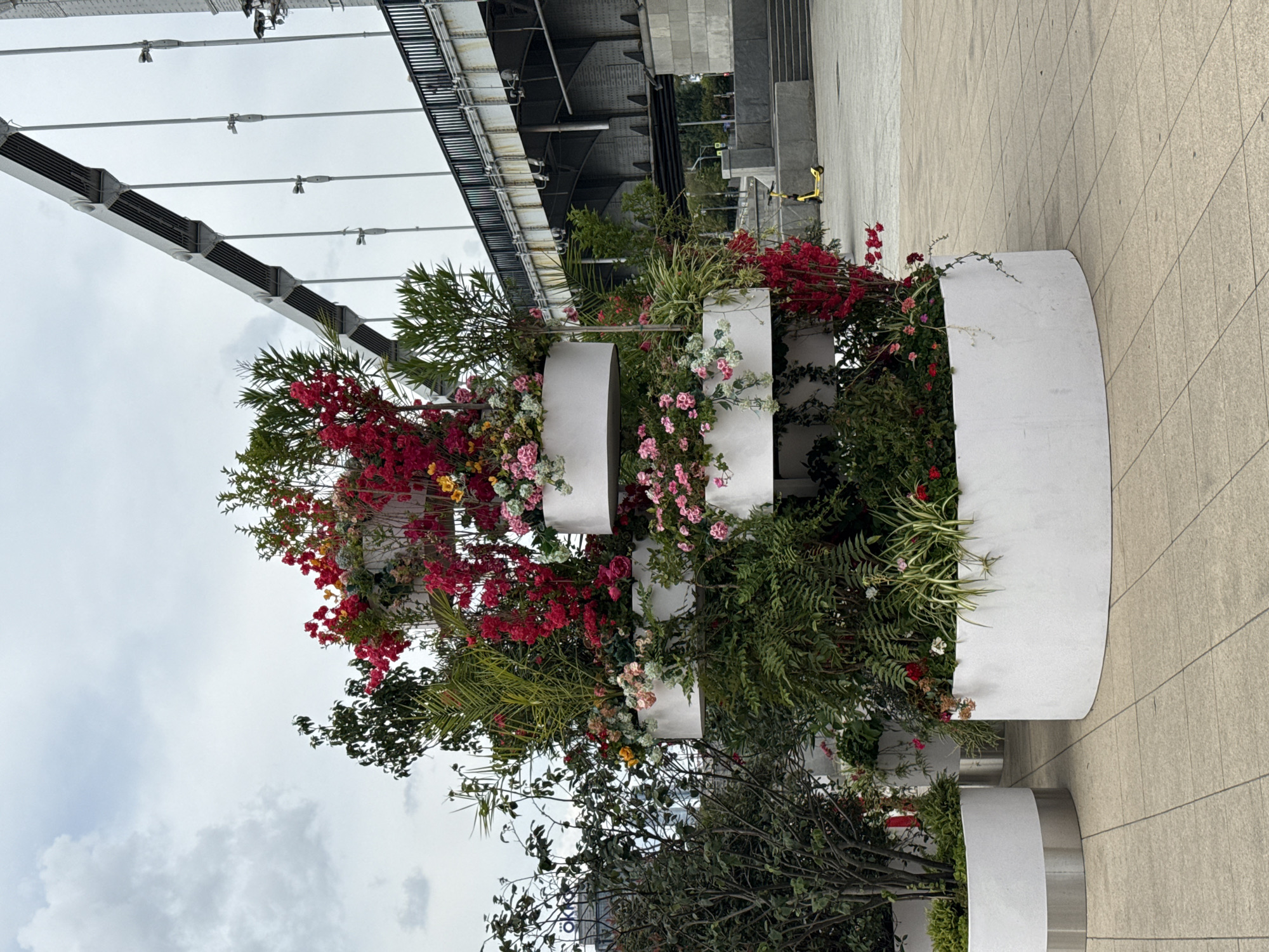 The image captures a large, multi-tiered planter filled with vibrant red, pink, and yellow flowers, as well as lush green foliage. The planter is situated on a paved outdoor area, likely a promenade or public square. In the background, a large suspension bridge with a metallic structure and cable supports dominates the skyline, suggesting an urban environment. A building with visible lettering, possibly "OKKO", is partially visible to the left. The sky is overcast with scattered clouds. To the right, a bright yellow electric scooter is parked near a set of concrete steps and a railing, indicating a public space with facilities for transportation and access. The overall scene suggests a well-maintained urban landscape, possibly near a river or waterfront, given the presence of the bridge. The location is likely Moscow, Russia, given the architectural style of the bridge and the visible building signage.