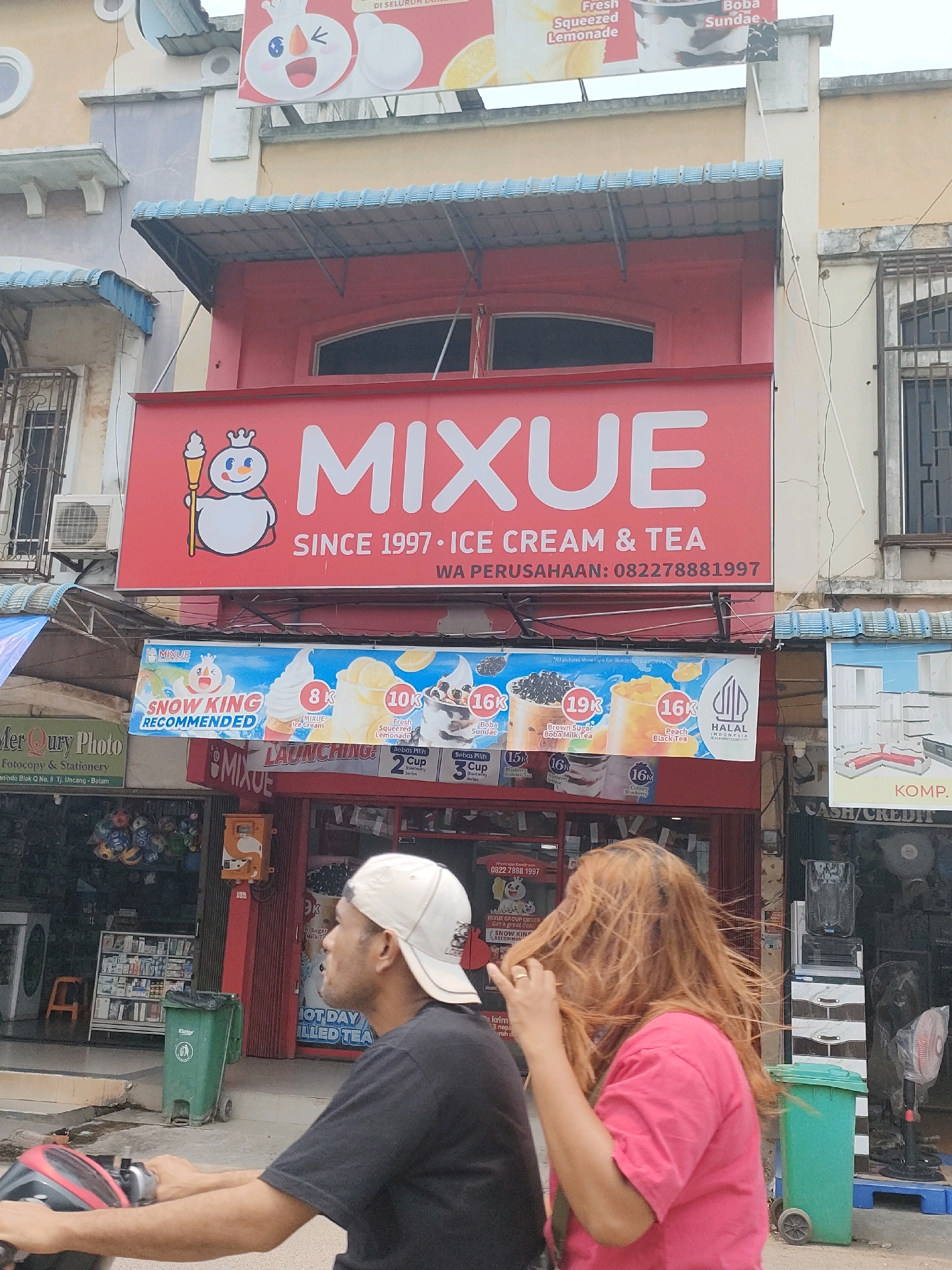 The image shows the exterior of a MIXUE store in Batam, Indonesia, likely in the Nagoya area based on the store's branding and typical urban streetscape. The store has a prominent red sign with the MIXUE logo and text that reads "MIXUE SINCE 1997 · ICE CREAM & TEA WA PERUSAHAAN: 082278881997". Below this sign is a banner advertising various MIXUE products, including "SNOW KING RECOMMENDED", ice cream, and drinks with prices indicated in thousands of Indonesian Rupiah (k).
In the foreground, two people are partially visible on a scooter. A man wearing a white baseball cap and a black t-shirt is driving. A woman with long, reddish-brown hair, wearing a pink t-shirt, is seated behind him, her hair blowing in the wind. They are moving away from the camera.
The street scene includes other shops and businesses. To the left of the MIXUE store, there is a shop labeled "Mer Qury Photo" which appears to be a photography and stationery shop. Various items are displayed in the windows of these shops, including what look like bottled beverages, stationery, and household appliances. Green trash bins are visible on the sidewalk. The buildings have multiple stories with visible windows and air conditioning units. The overall environment suggests a commercial street in an urban setting.