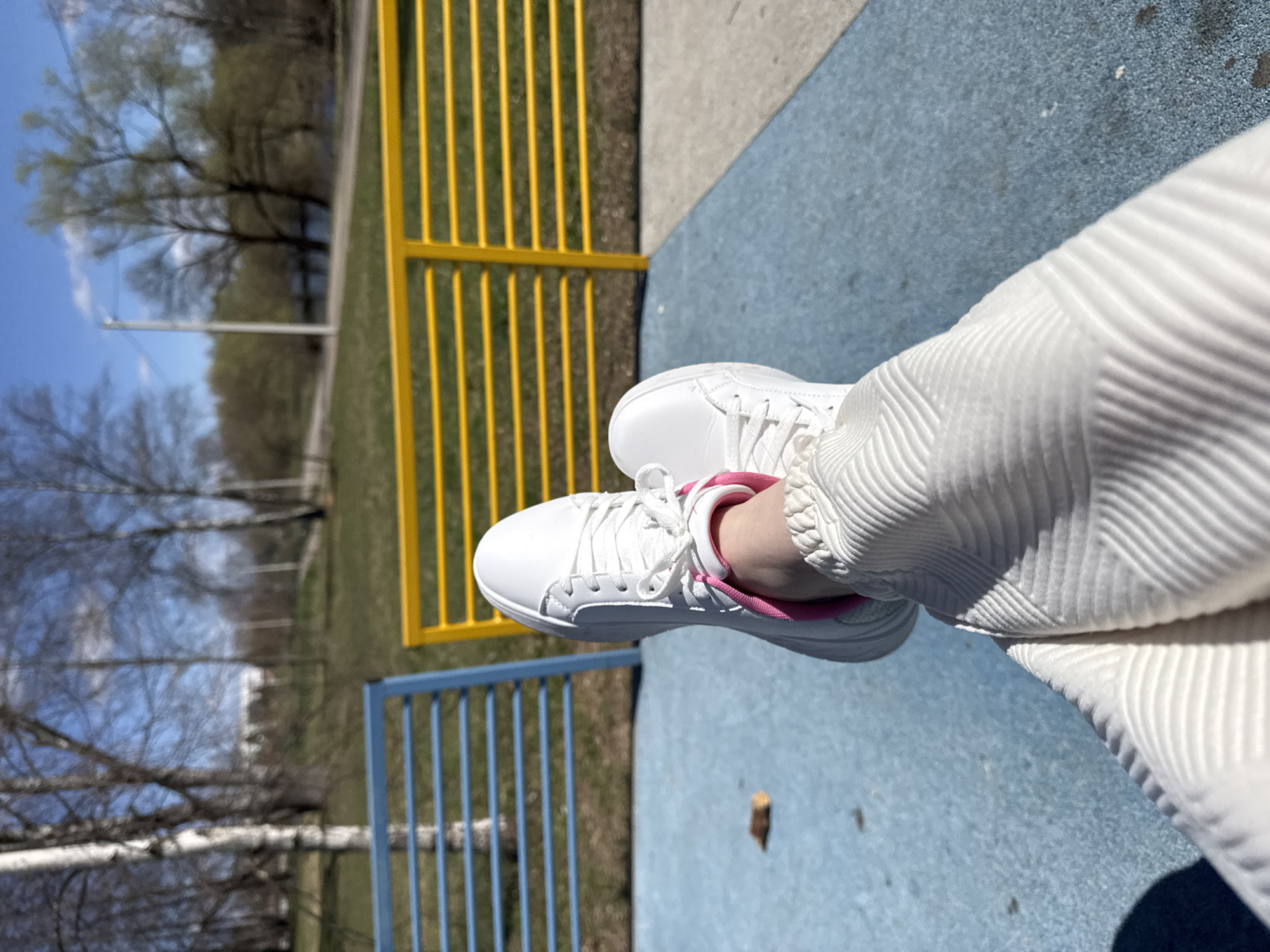 The image shows a person's legs and feet in white clothing and white sneakers with pink accents. They are sitting on a blue surface with a yellow and blue metal fence in the background. The background shows trees and a grassy area, suggesting an outdoor park setting. While the location is stated to be Shevlyakovo, Russia, there are no clear identifying markers to confirm this without additional context or information.