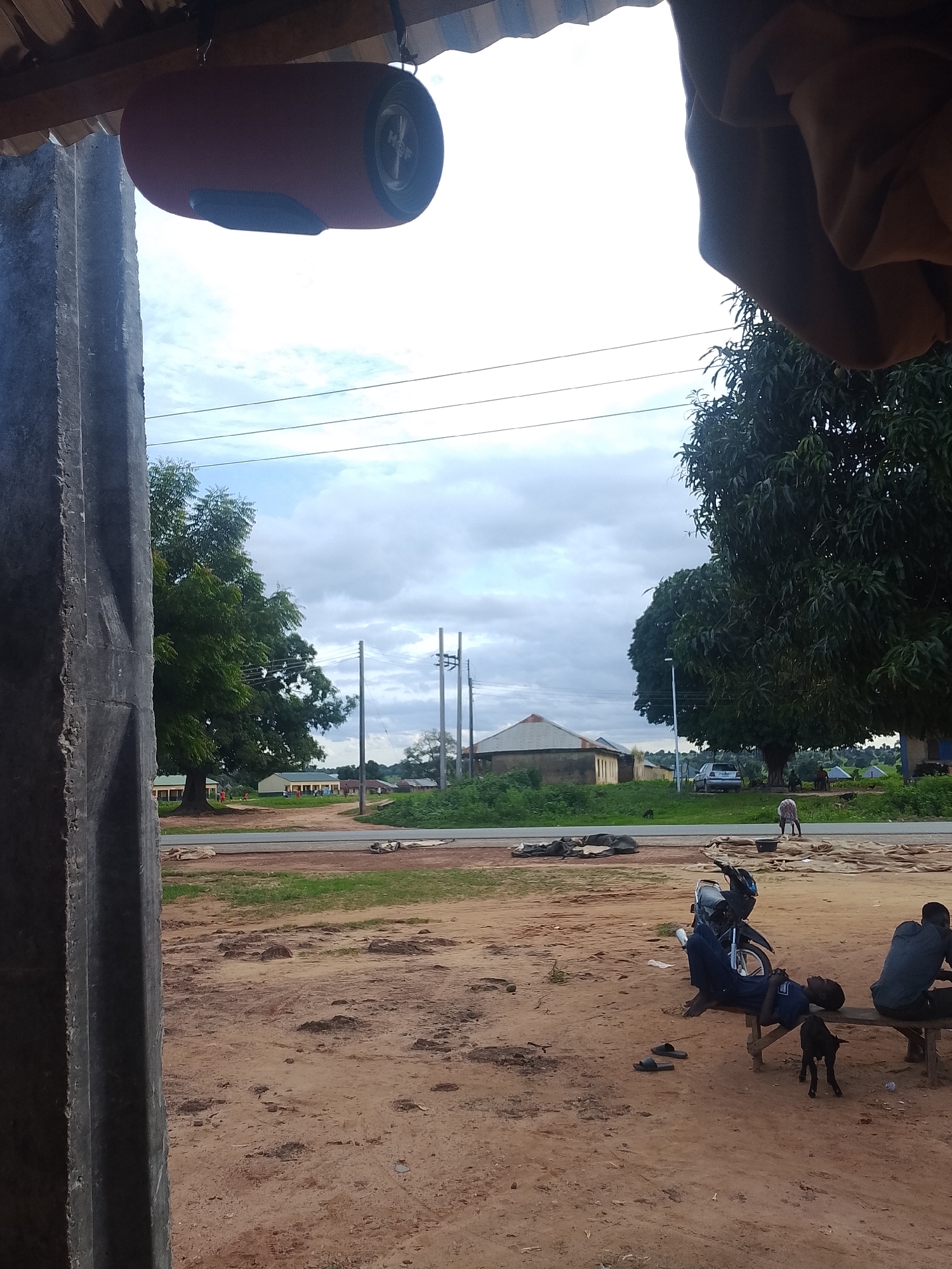 A ground-level outdoor shot captures a dirt road and a sandy clearing, with a concrete pillar in the foreground on the left. The sky is overcast with clouds. In the middle ground, a motorcycle is parked, and two young men are seated on a wooden bench. A small black dog stands between them. To the right of the motorcycle, another person is visible, and further back, a car is parked. In the background, several buildings with white walls and light-colored roofs are present. A tall tree with dense green foliage stands prominently behind the motorcycle and the bench. A few other smaller trees and bushes are scattered throughout the scene. In the far background, a large, dark green tree dominates the view. Overhead, a red cylindrical object, possibly a speaker, hangs from a beam.