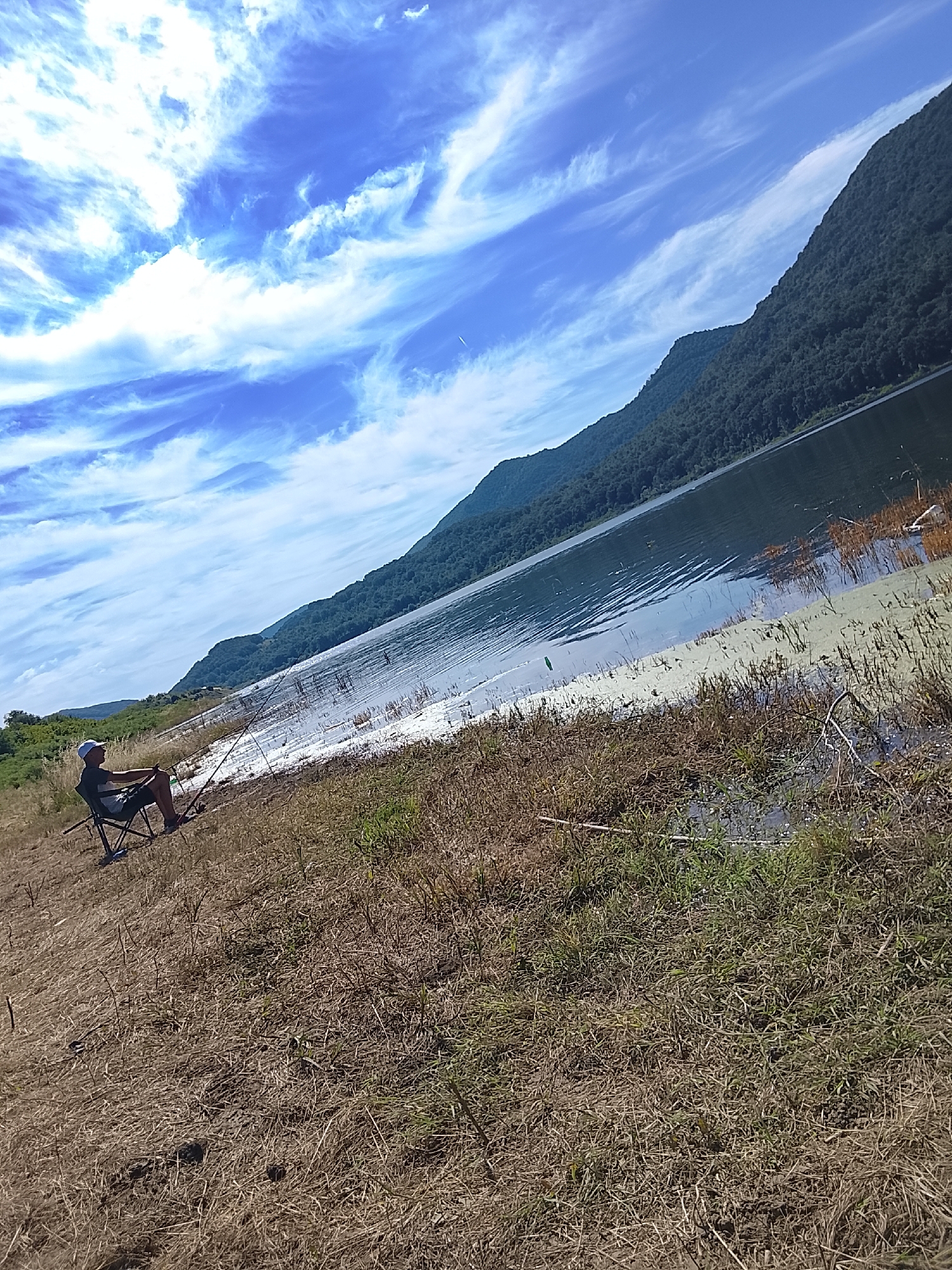 The image depicts a serene scene by a lake, likely in Asparuhovo, Bulgaria. The main subject is a person sitting in a folding chair on the grassy bank, holding a fishing rod, engrossed in the activity of fishing. The person is wearing a blue cap, a dark t-shirt, and shorts. A fishing rod is extended towards the water, suggesting an ongoing attempt to catch fish.
The setting is a natural landscape with a vast lake reflecting the sky and the surrounding forested mountains. The mountains, covered in dense green foliage, rise majestically on the far side of the lake, creating a dramatic backdrop. The sky is a vibrant blue, adorned with wispy, white clouds that streak across it, adding a sense of movement and depth. The weather appears to be fair and sunny, typical of a pleasant day. The time of day is likely late morning or early afternoon, judging by the bright sunlight.
The foreground is dominated by dry, yellowish grass and some scattered green patches, indicating a semi-arid environment or late summer season. The lake's edge shows some aquatic vegetation and possibly some debris. The overall mood is peaceful and tranquil, evoking a sense of calm and solitude. The gentle ripples on the water's surface suggest a light breeze. There is no visible text in the image.
