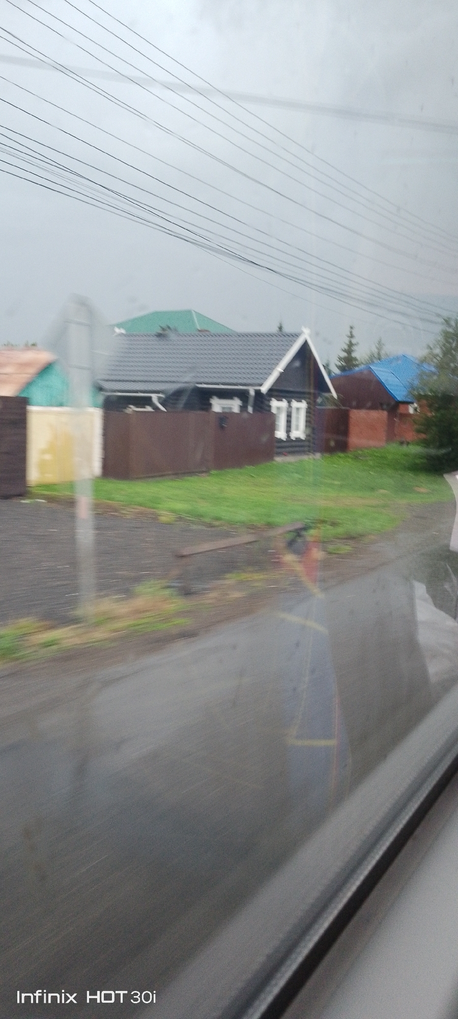 The image depicts a row of houses with varying colored roofs, situated in what appears to be a residential area. The houses are of traditional construction, with one house featuring a dark-colored wooden exterior and white trim around the windows. Another house has a light blue roof, and one has a green roof. A brown wooden fence runs along the front of the properties. The ground in front of the houses is covered with green grass. The scene is set against a cloudy sky, and there are power lines running across the top of the frame. The road in front of the houses appears wet, suggesting recent rain. The text "Infinix HOT 30i" is visible in the bottom left corner, indicating the device used to capture the image. The location is identified as Yemelyanovo, Russia.