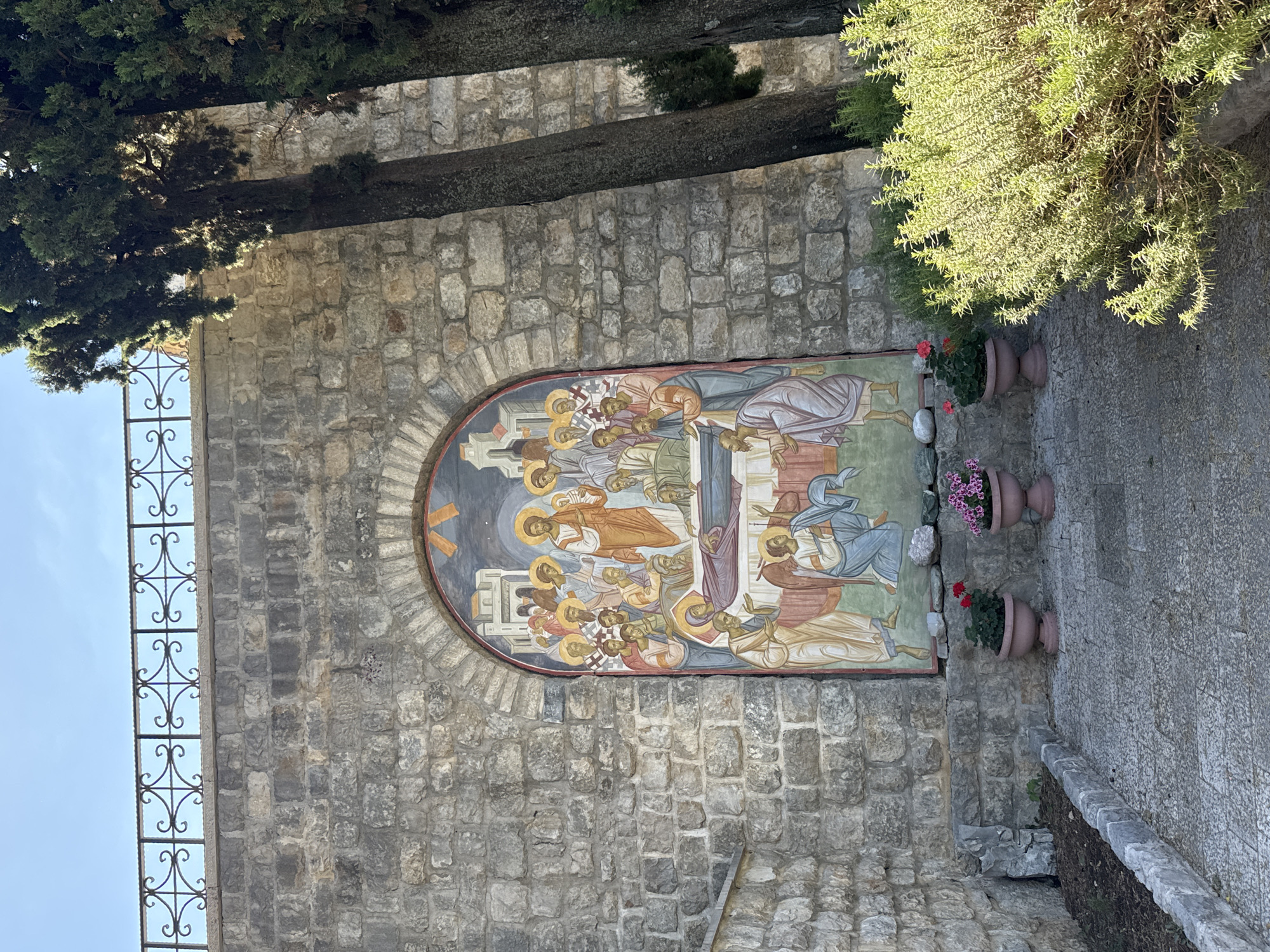 The image depicts an outdoor mural on a stone wall in Trebinje, Bosnia and Herzegovina, likely within the grounds of a monastery or church. The mural, painted in a traditional Byzantine style, portrays a religious scene with figures in flowing robes and halos. In the center, Christ is depicted, surrounded by other figures who appear to be mourners or disciples. A central element of the mural shows the body of Christ being laid to rest. The mural is set within an arched recess in the stone wall. In front of the mural, a paved walkway is visible, leading to a small garden area with potted plants, including red and pink flowers, and a lush green shrub. The stone wall is constructed from rough-hewn blocks, and a decorative wrought-iron railing runs along the top. Tall trees with dark bark and dense foliage are situated to the right of the mural, casting shadows on the scene. The sky above is clear and blue.