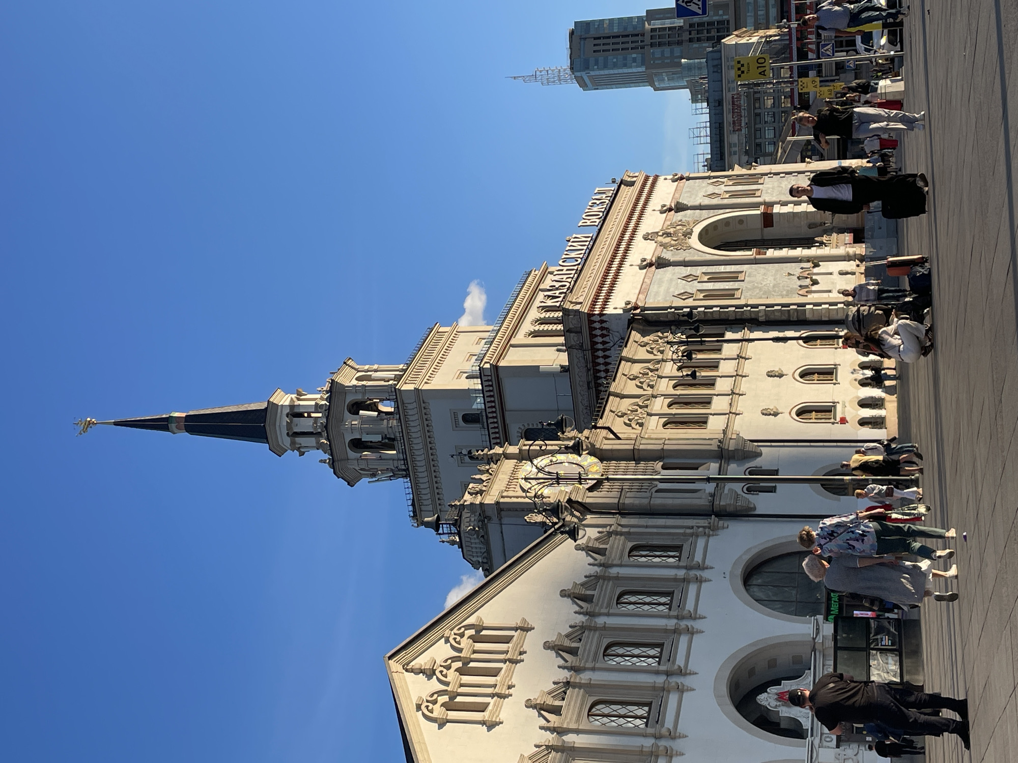 This media file captures a bright, sunny day in Moscow, Russia, with a clear blue sky dominating the upper portion of the image. The primary subject is the Kazan Railway Station, a grand architectural landmark characterized by its ornate facade, multiple windows, and a prominent spire topped with a golden weathervane. The building exhibits classical architectural elements with intricate detailing and decorative moldings.
In the foreground and mid-ground, several people are seen in the plaza in front of the station. They appear to be travelers or visitors, some with luggage, suggesting the setting is a busy transportation hub. The people are casually dressed, indicating a pleasant daytime temperature. Their postures and interactions do not convey any specific strong emotions, but rather a sense of ordinary activity, such as walking, waiting, or conversing.
Visible text on the building includes "КАЗАНСКИЙ ВОКЗАЛ" (Kazan Railway Station) written in large white letters on the facade. Other signs and text are visible on distant buildings, including what appears to be taxi signage ("A10", "A9"), and a green sign on the station's lower level that reads "МЕГАФОН" (Megafon), a telecommunications company. In the background, modern skyscrapers are under construction, providing a contrast between historical and contemporary architecture in Moscow. The overall scene depicts a typical day at a major city train station, bustling with activity under a clear sky. The time of day appears to be midday, given the strong sunlight and the shadows cast.