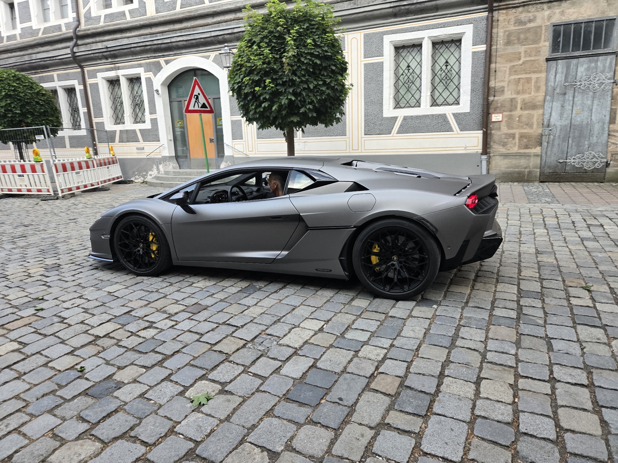 A grey Lamborghini Aventador is driving on a cobblestone street in front of a building with a stone facade and a grey-and-white patterned wall. The building has arched doorways and windows with ornate grilles. A round, green tree stands to the right of the car, and a temporary construction barrier with orange and white stripes is visible on the left. A traffic sign depicting a shovel and a pile of dirt is positioned near the building's entrance. The car's driver, a man with a light-colored shirt and sunglasses, is visible through the window. The scene appears to be in a historic district, likely in Ansbach, Germany, given the architectural style.
