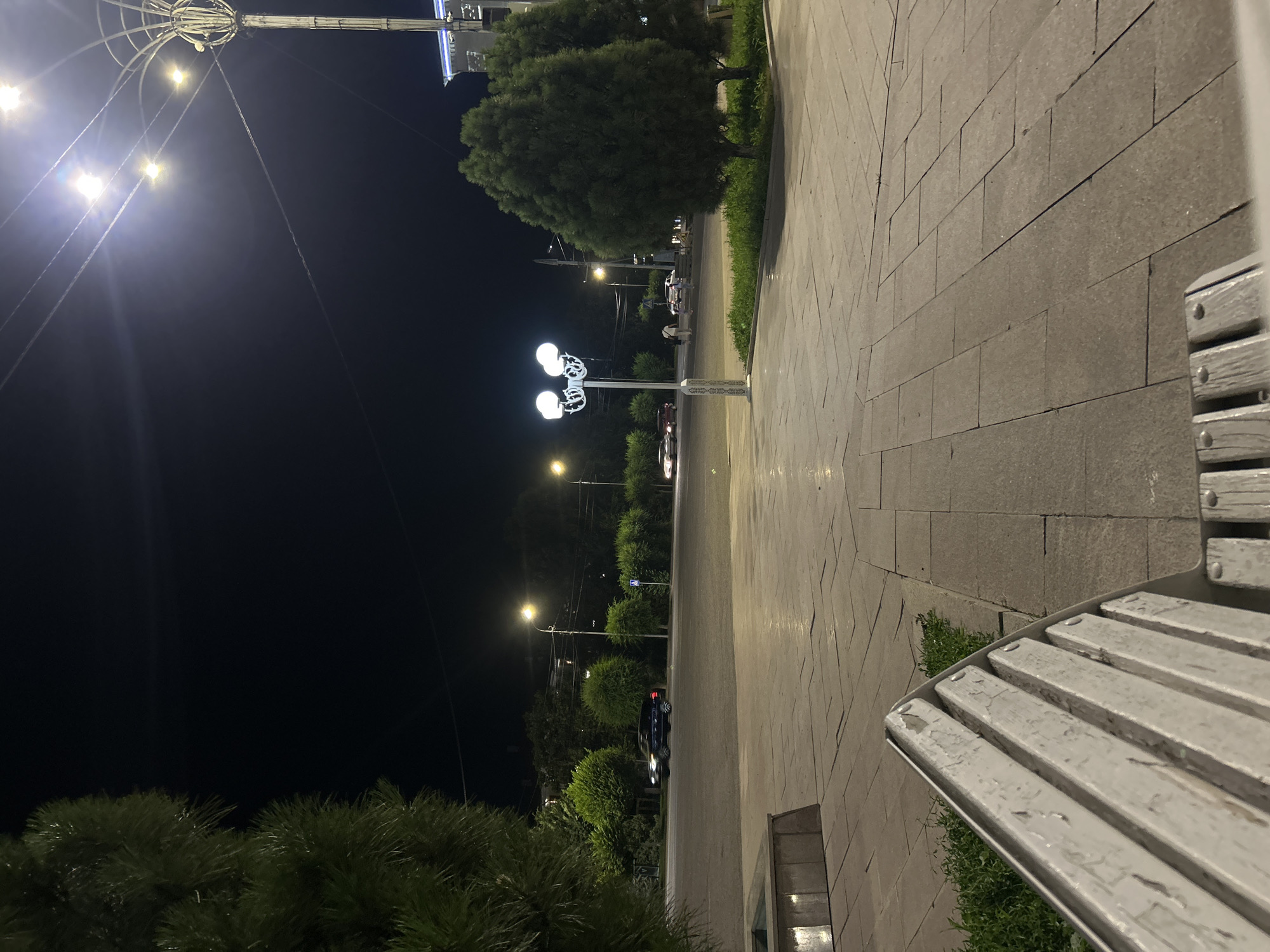 This image captures a nighttime scene on a paved street in the city of Taraz, Kazakhstan, near the city administration building. In the foreground, the edge of a white wooden bench with peeling paint is visible. The ground is paved with large, light-colored square tiles, reflecting some of the ambient light. To the left, the dark, lush foliage of a pine tree partially obscures the view.
The middle ground shows a street with active traffic. Several cars are visible, including a dark SUV driving away from the camera and two cars in the distance, one red and one white, their headlights illuminated. On the right side of the street, a decorative lamp post with multiple globes illuminates the area. Behind the lamp post and across the street, the dark silhouettes of trees and buildings can be seen. Further down the street, another lamp post with two globes stands. Several individuals are walking on the sidewalk across the street, their figures indistinct in the low light. The sky is dark, indicating it is night, with some light pollution from the city visible, particularly around the streetlights.