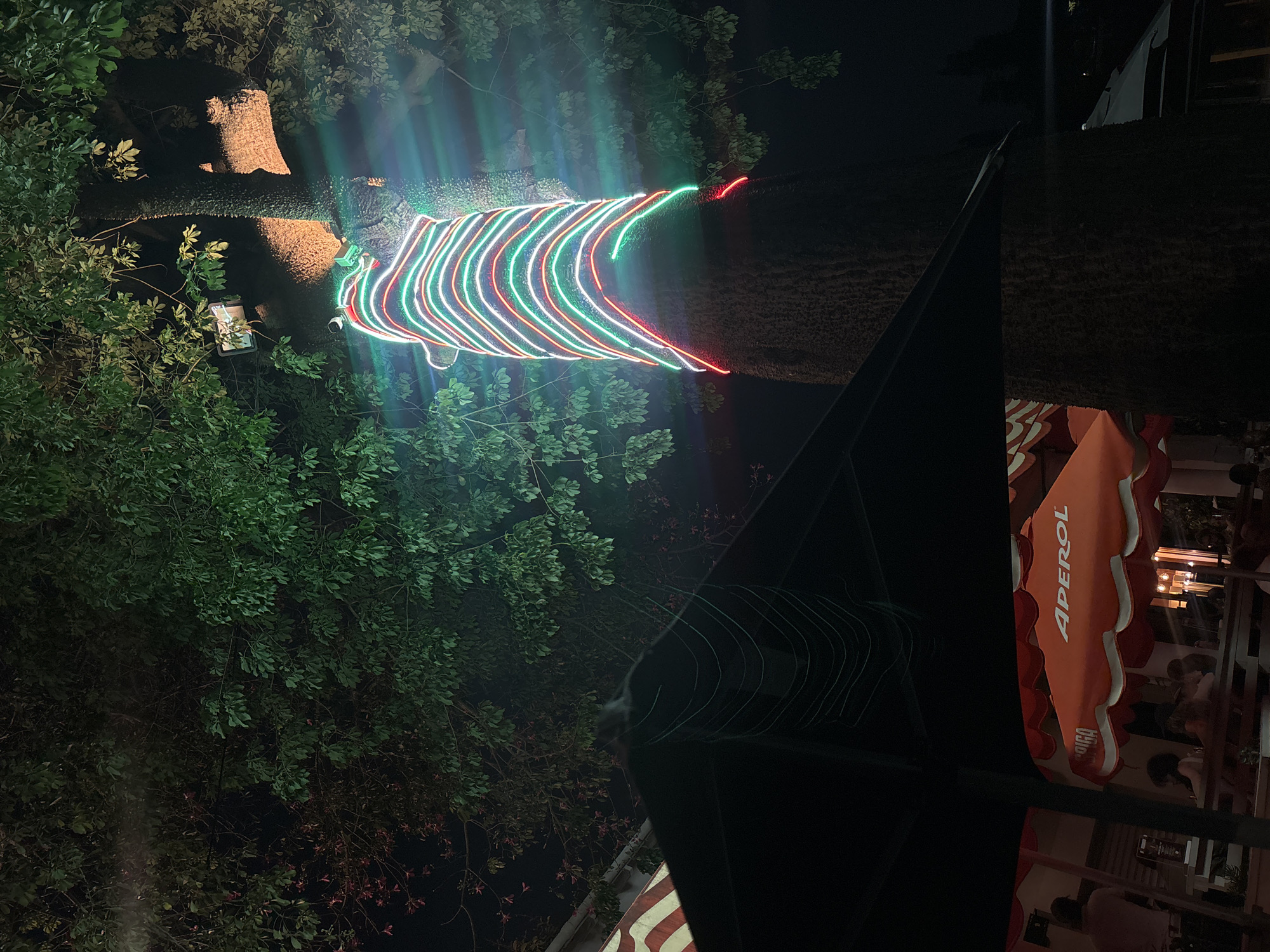 Here is a description of the image:
At night, a tree is decorated with strings of red, white and green lights. The tree is surrounded by foliage and a black canopy. To the right, an Aperol umbrella is visible, and people can be seen inside a building. The scene is most likely at a bar or restaurant in Durban, South Africa.