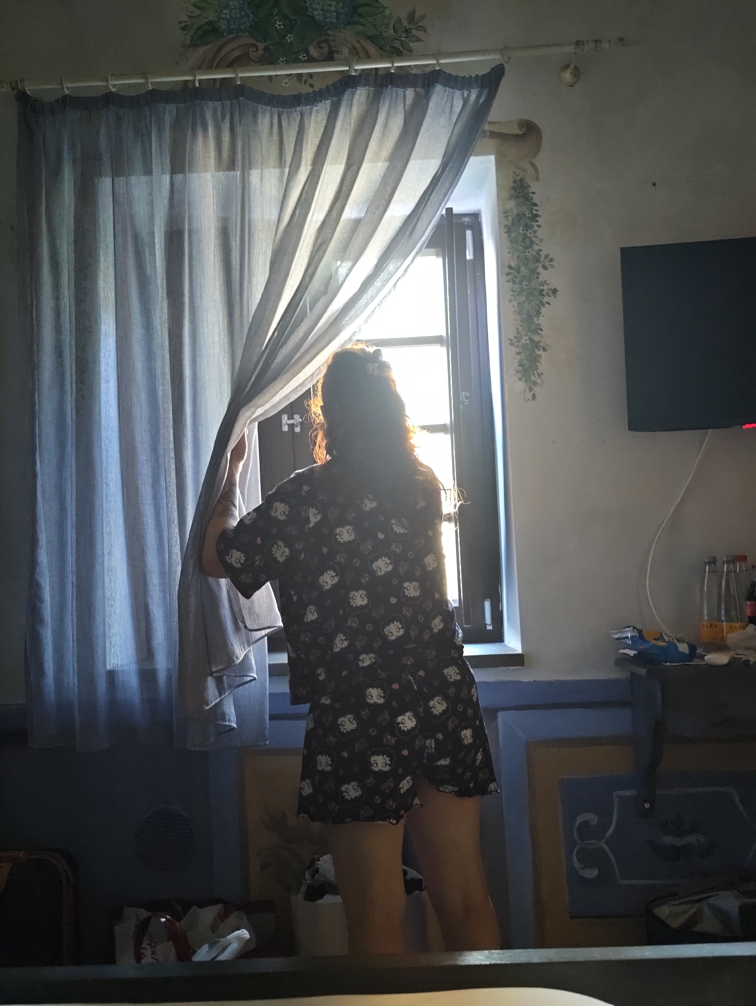 A woman with long, wavy, reddish-brown hair stands in a dimly lit room, partially obscured by a sheer gray curtain she is holding open. She is wearing a dark short-sleeved dress or top with a repetitive pattern of cartoon faces. She is looking out a window where bright sunlight is streaming in, silhouetting her. The room has textured walls with painted decorative elements, including a floral motif above the curtain rod and a trailing vine pattern on the right wall. A dark television is mounted on the right wall. Below the window, a blue and yellow decorative trim runs along the wall. On a surface below the television, there are several bottles and some crumpled packaging. In the foreground, a portion of a dark piece of furniture is visible, with some items on it. The overall impression is that of an indoor scene, possibly a bedroom or living space, with the natural light from the window being a significant element. The image does not provide any specific visual cues to pinpoint the exact location within the city of Pozzuoli, Italy.
