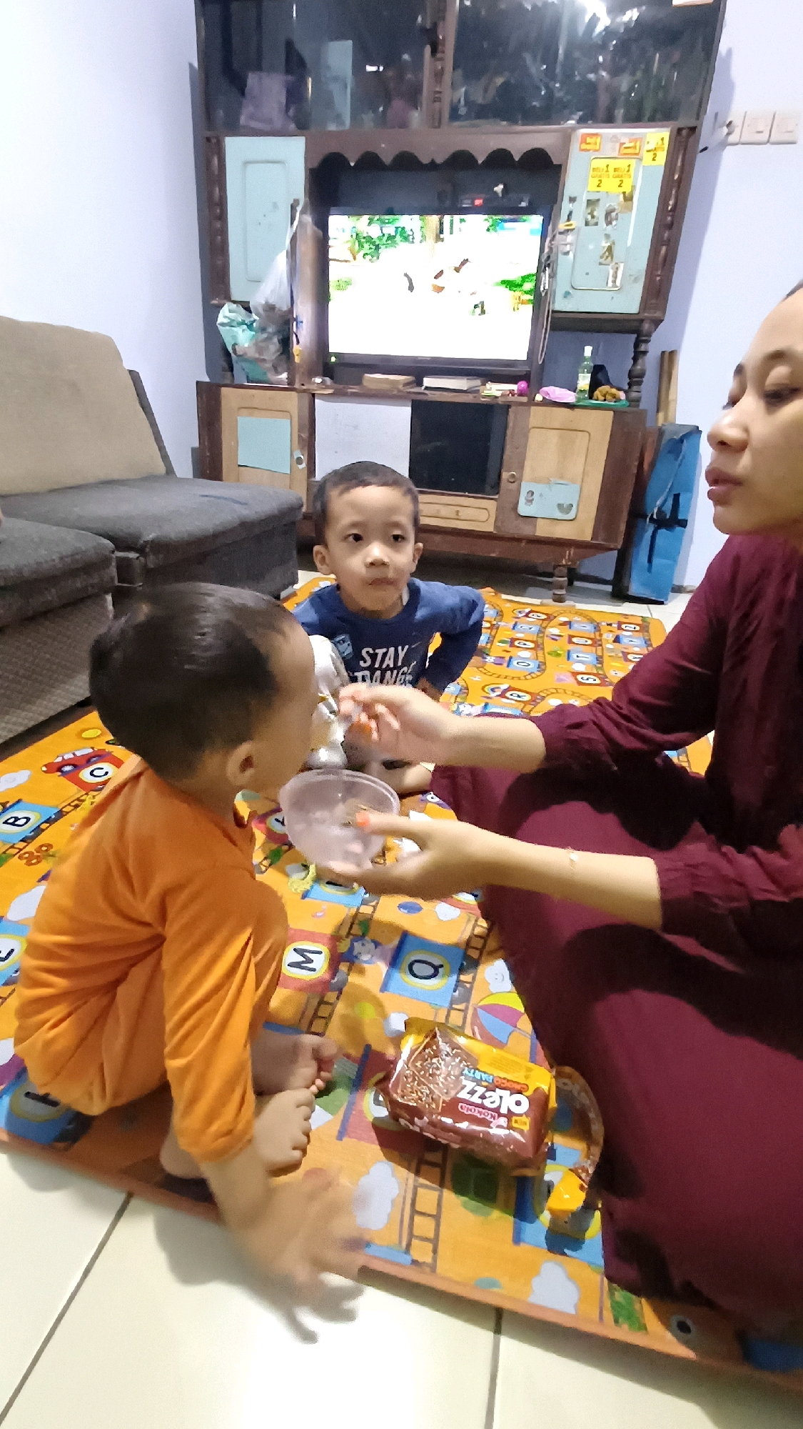 In Kowangan, Indonesia, a mother is feeding her young child from a small clear bowl. The child, wearing an orange shirt, is sitting on a colorful mat with cartoon characters and alphabet letters. A second child, in a blue shirt, watches nearby. A packet of chocolate wafers sits on the mat, and a television is visible in the background. The setting appears to be a living room.
