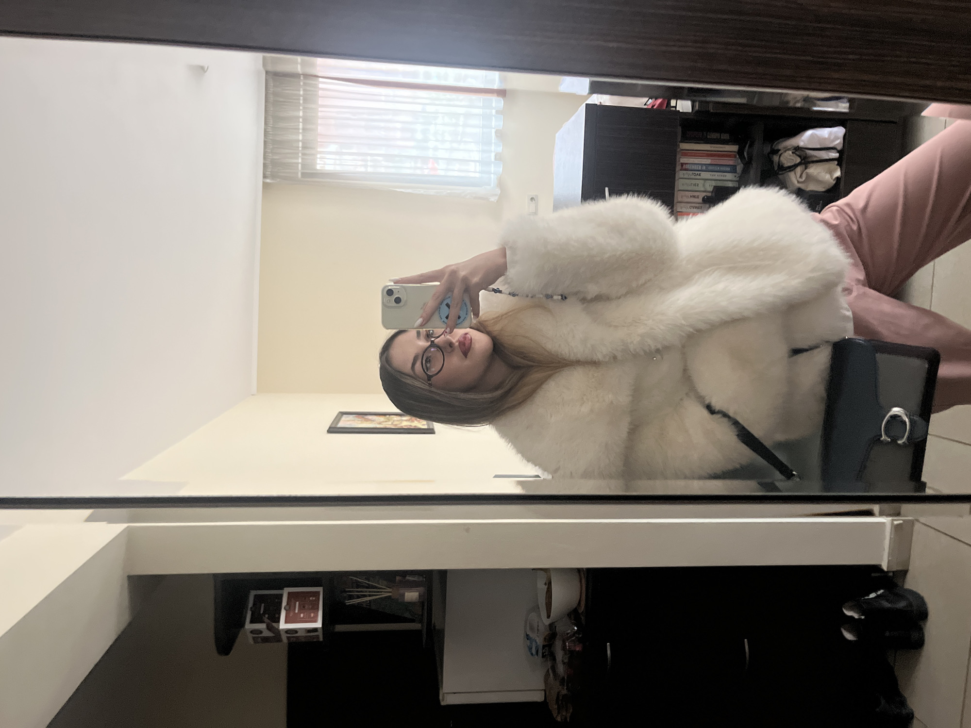 This mirror selfie captures a young woman indoors, likely in Sofia, Bulgaria. The image, presented with a 90-degree clockwise rotation, shows the woman taking her picture using a light-colored smartphone with a circular emblem on its back.
The subject is a woman with long, light-colored hair, wearing round-framed glasses and dark red lipstick. She is dressed in a voluminous, white, fluffy faux fur coat and light pink pants. She holds a dark handbag with a silver ring clasp on her right arm. Her gaze is directed towards the phone, indicating she is posing for the photo.
The background, reflected in the mirror, reveals a well-lit interior space. Natural light enters from a window fitted with white Venetian blinds in the upper right. A dark wooden shelving unit is visible to the woman's right, filled with stacked books and a bundled white cloth. On the left side of the reflection, a light-colored wall displays a small framed picture, and below it, a small white appliance, a white mug, and a diffuser are visible on a surface. The walls of the room are light-colored.
The scene depicts a personal moment of self-photography, likely showcasing an outfit. The presence of natural light suggests it is daytime. No specific text on the books or the phone's sticker is clearly decipherable. The overall atmosphere is casual and domestic.