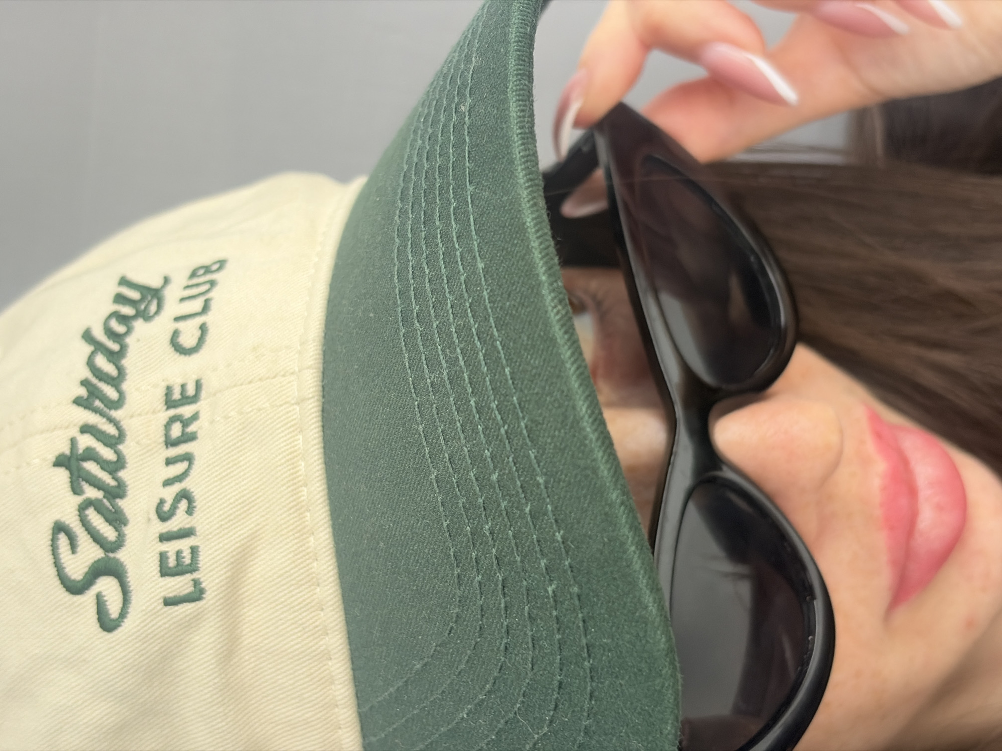 The image presents a close-up, cropped view of an individual's face, specifically from the nose downward, along with a portion of their right hand, against a plain, light grey background. The individual is wearing a two-toned baseball cap featuring a light beige crown and a dark green brim. The crown is embroidered with the text "Saturday" in green script, above "LEISURE CLUB" in green block letters. Black, cat-eye style sunglasses are worn, obscuring the eyes, with the cap's brim casting a shadow further downward. The visible lower portion of the face displays lips colored with a pink or reddish-pink lip product. The person's right hand, characterized by long, manicured fingernails with a white French tip design, is visible on the right side of the frame, in the process of touching or adjusting the right temple of the sunglasses. Dark brown hair is visible along the right side of the frame, extending downward. The scene is described as being in Saint Petersburg, Russia.