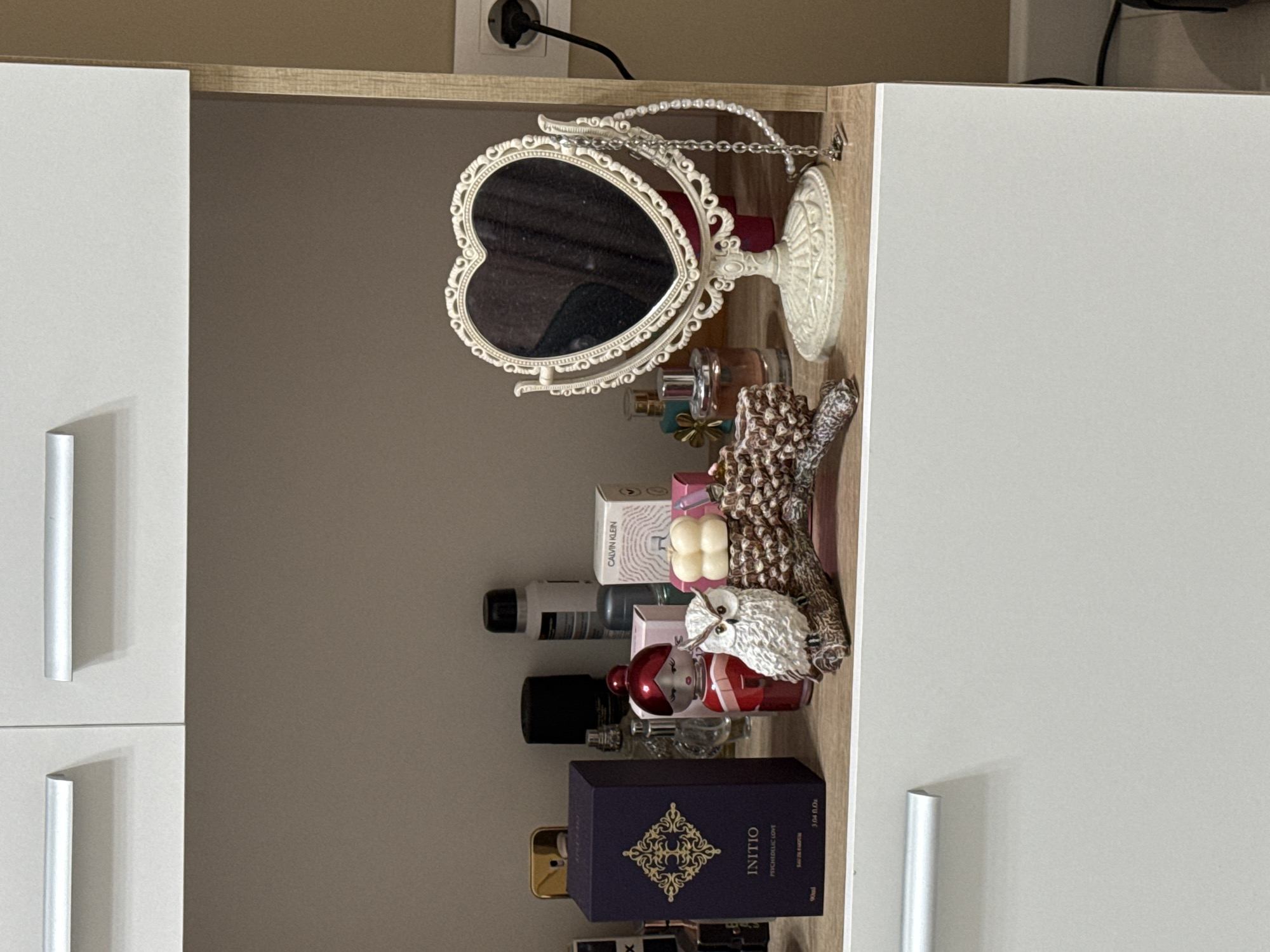 The image shows the interior of a cabinet or closet, likely in a residential setting in an unknown city. Inside, there is a collection of personal care items and decorative objects. Prominently featured is a heart-shaped mirror with an ornate white frame, positioned at the top. Below the mirror are various perfume bottles, including one with the brand name "INITIO" visible on a dark purple box. Other items include a Calvin Klein box and a decorative owl figurine. The cabinet has white doors with long, silver handles on the left side of the frame. A power outlet and cord are visible at the top. The environment appears to be organized and clean.