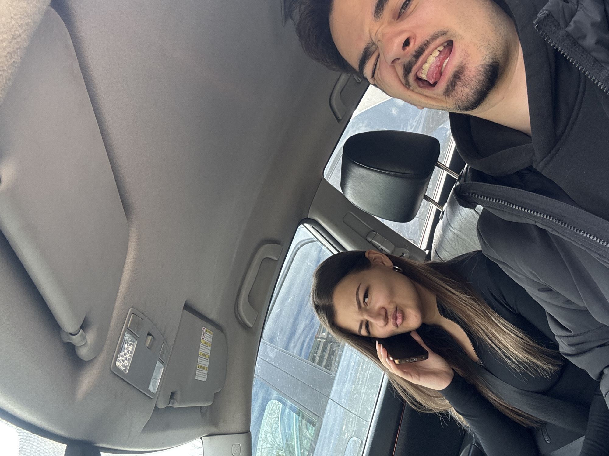 Two people are in a car. A man is in the front passenger seat wearing a black jacket and making a face. A woman is in the driver's seat, wearing a black outfit, talking on her mobile phone, and making a kissing face. The interior of the car is visible, including the dashboard, sun visor, and seats. The exterior visible through the windows shows what appears to be a parked car and a building. It's difficult to determine the location in Sofia, Bulgaria, specifically without further landmarks or context.