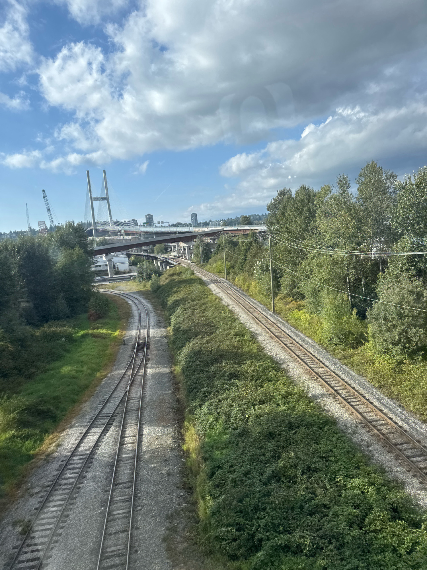 This is an eye-level, high-angle, wide shot of a train track, in Surrey, Canada, as viewed from a window. The train track has two sets of rails that curve to the left and then to the right, disappearing into the distance. On the left side of the tracks, there is a grassy embankment. On the right side of the tracks, there is a dense forest of green trees, with power lines running through them. In the background, there is a large cable-stayed bridge with two tall towers, and beyond that, a cityscape with buildings. The sky is blue with white and gray clouds.