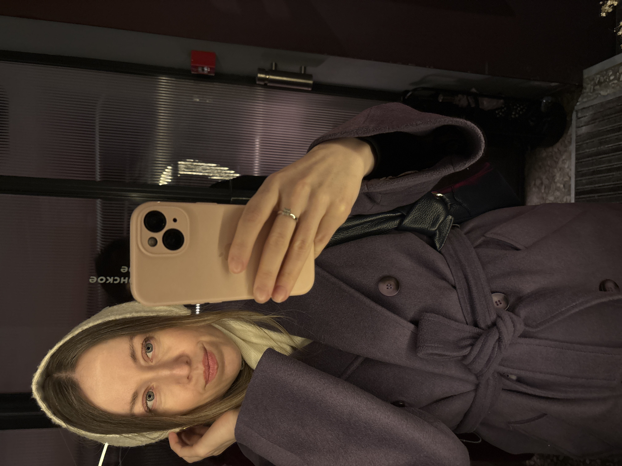 A woman wearing a white hood and a plum-colored coat is taking a selfie. She is holding a peach-colored phone. The background appears to be an elevator or public transportation space. The words "HCKOE" are seen reflected in the background. This text suggests that the location is in Saint Petersburg, Russia, as that word is a part of station names.