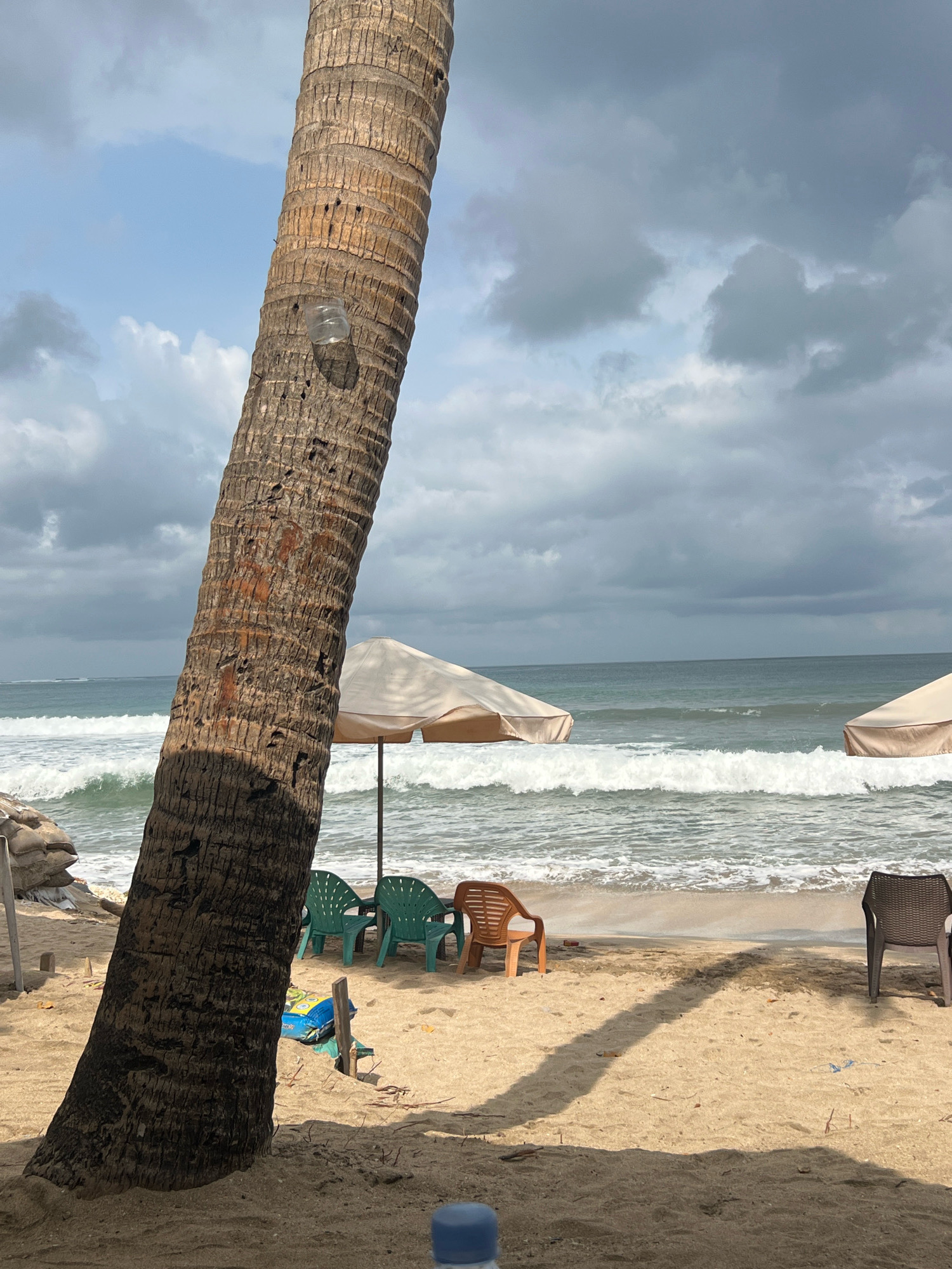 The image depicts a beach scene with a prominent palm tree trunk in the foreground. Behind the tree, the ocean with rolling waves is visible under a cloudy sky. Several beach chairs and umbrellas are set up on the sandy shore. On the left side, there are sandbags. The overall impression is a somewhat overcast day at a beach that appears to be in or around Kuta, Indonesia, a popular tourist destination known for its beaches.